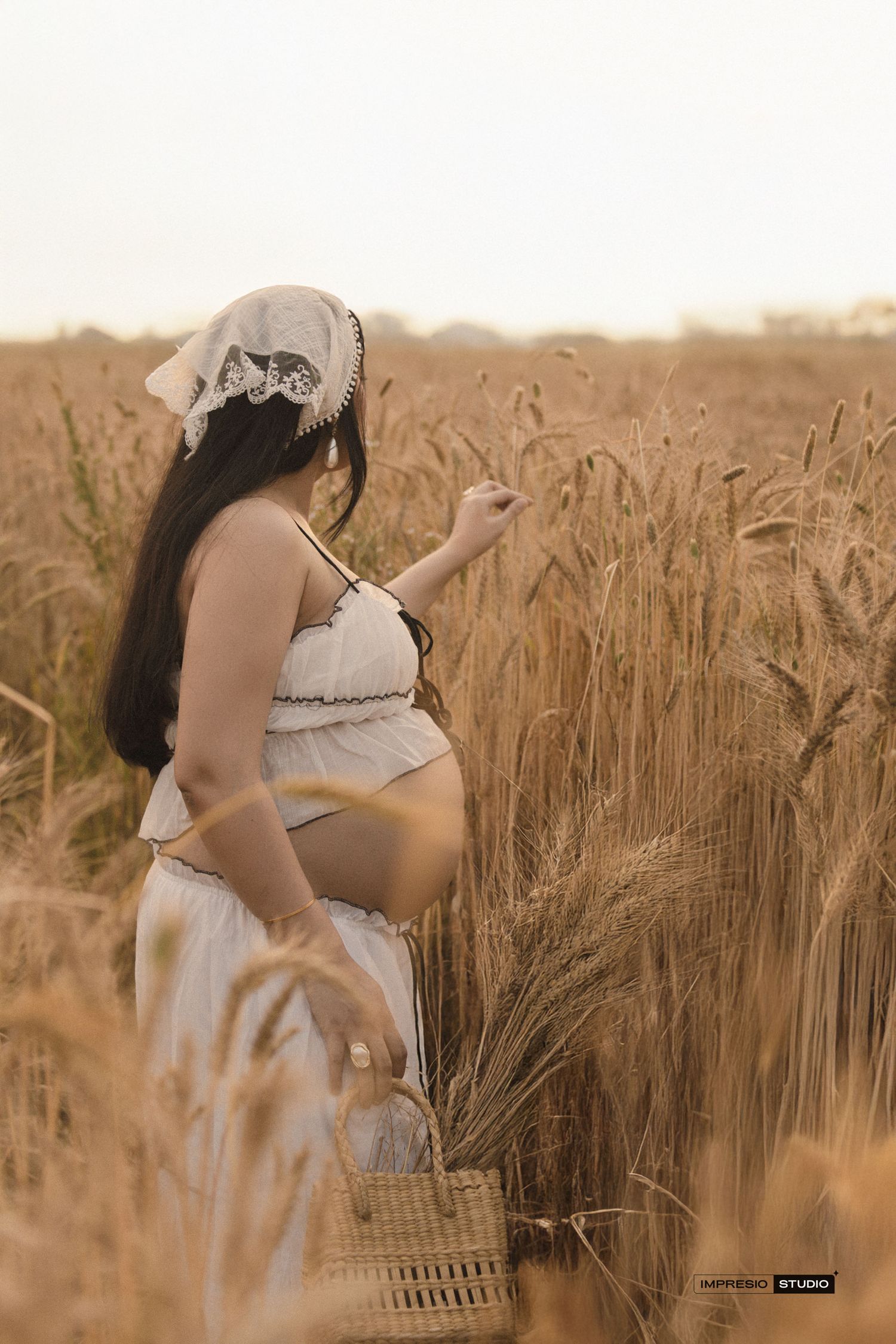A pregnant woman in white stands in a golden wheat field holding a wicker basket at sunset.