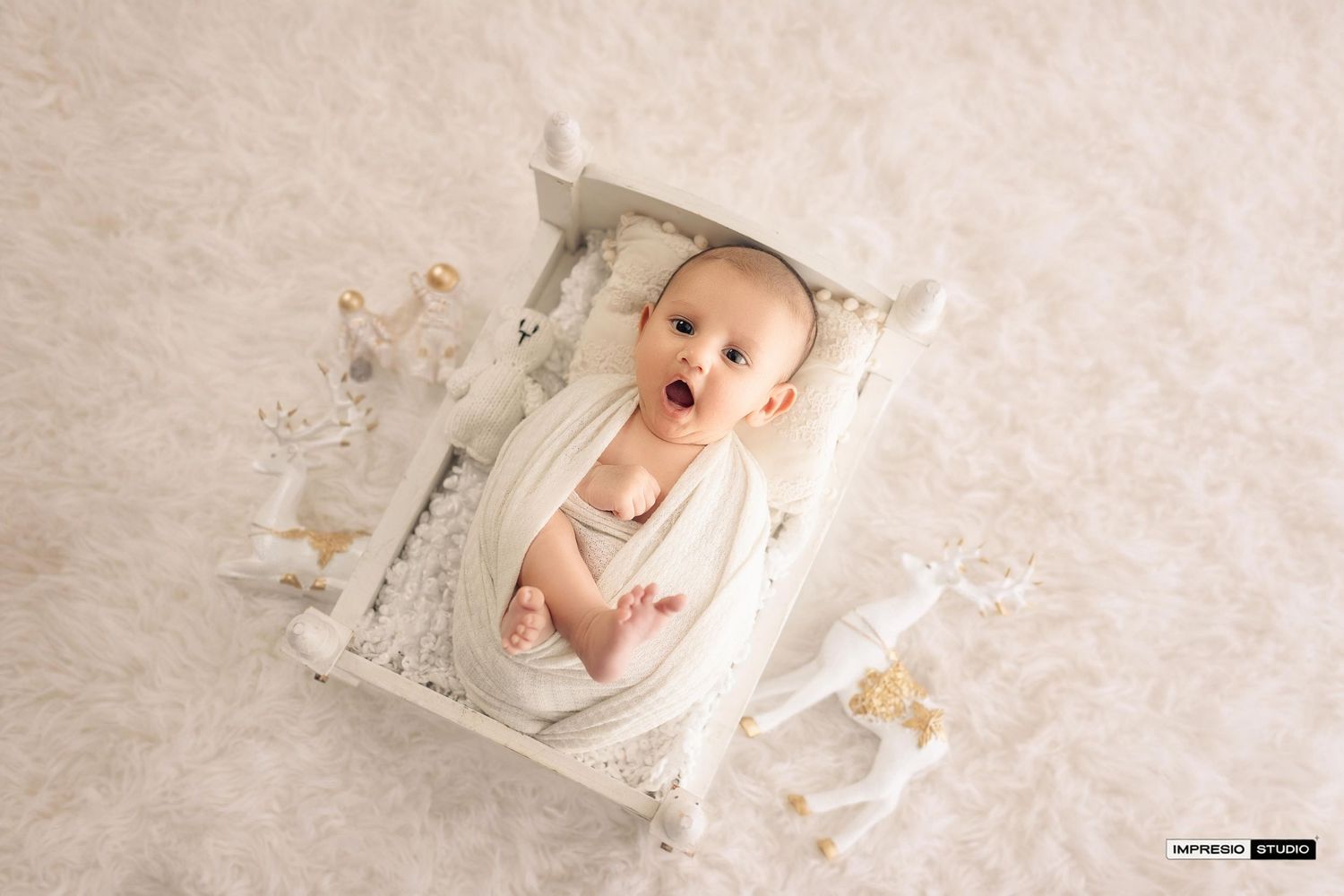 A wide-eyed baby wrapped in white cloth lies in a small white wooden bed surrounded by delicate white decor.