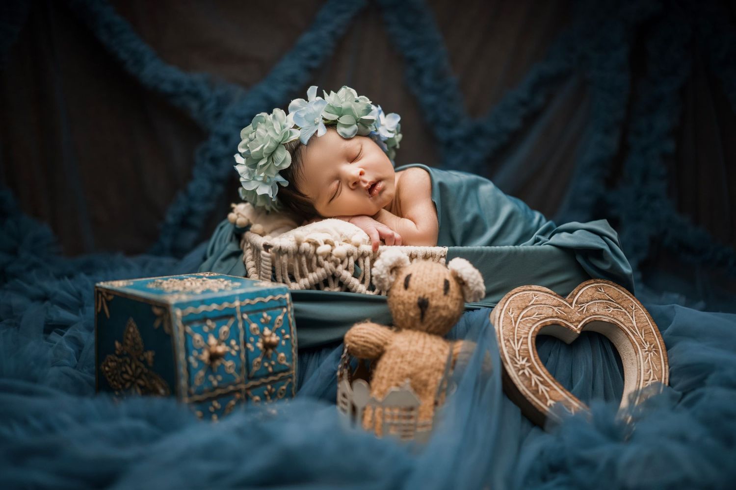 Sleeping newborn baby wearing floral crown in blue basket surrounded by teddy bear and decorative props.