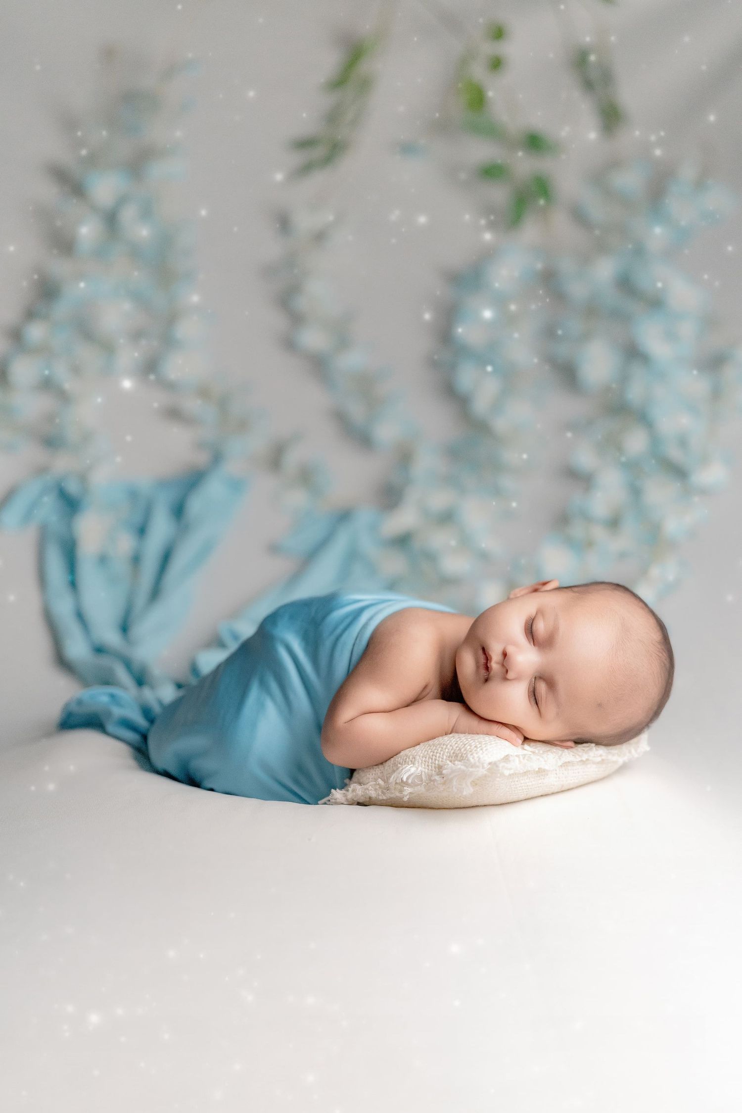 Newborn baby sleeping peacefully wrapped in blue fabric on a small pillow with a soft floral backdrop.