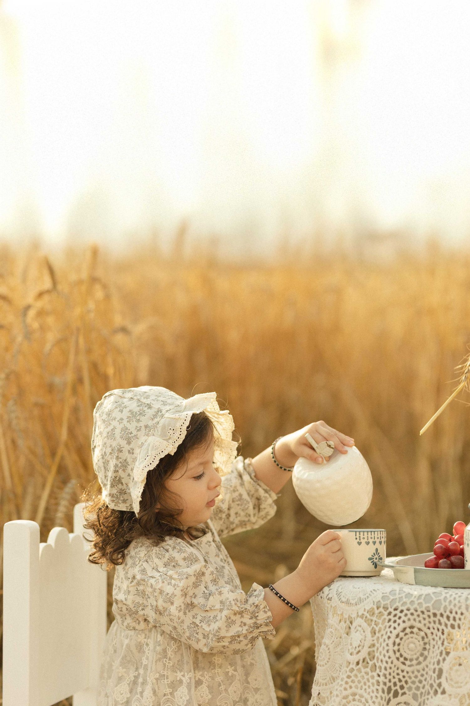 Young girl in floral dress and bonnet pouring tea at a lace-covered table in a golden wheat field.