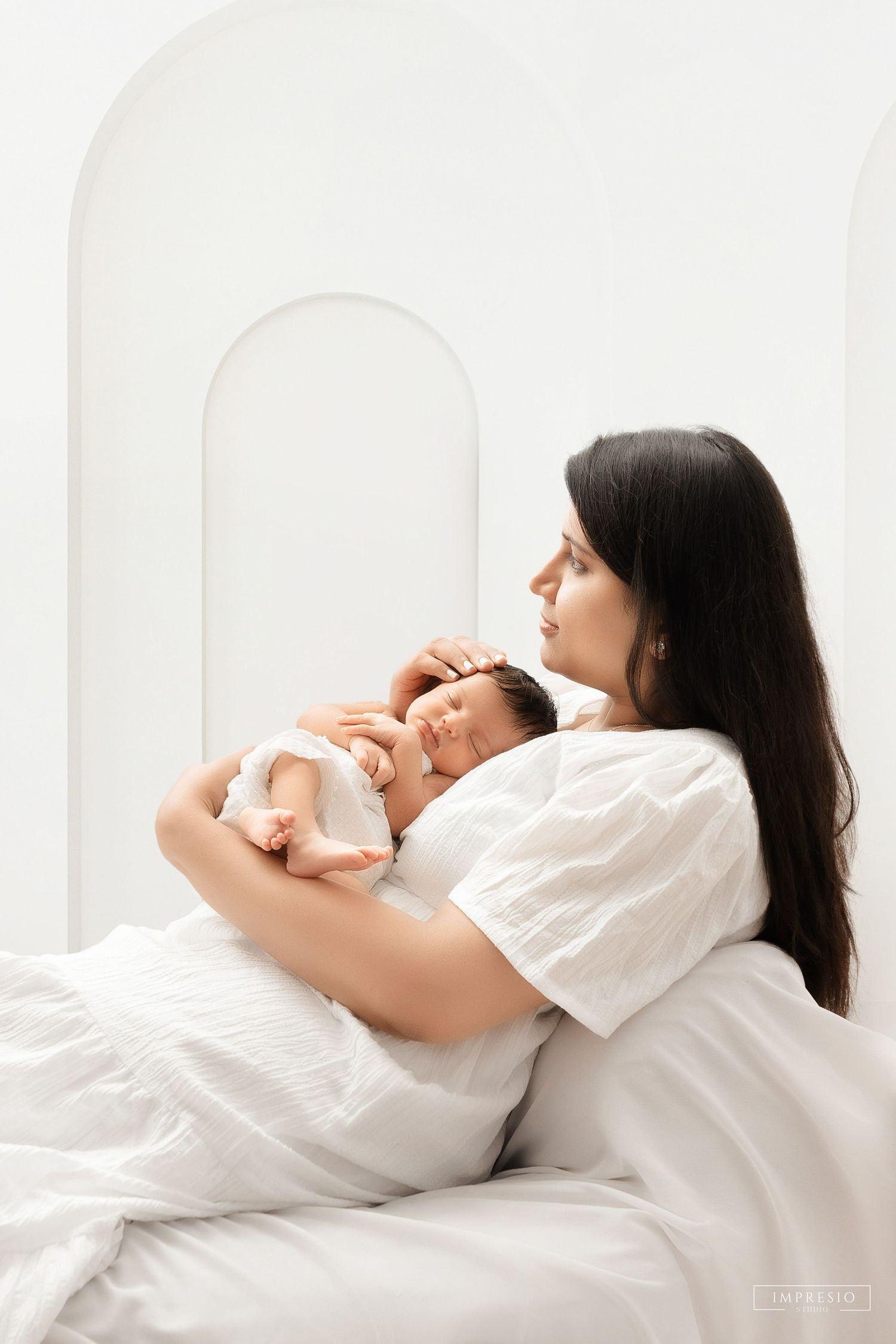 Mother in white dress lying on bed gently cradling her sleeping newborn baby against white backdrop.