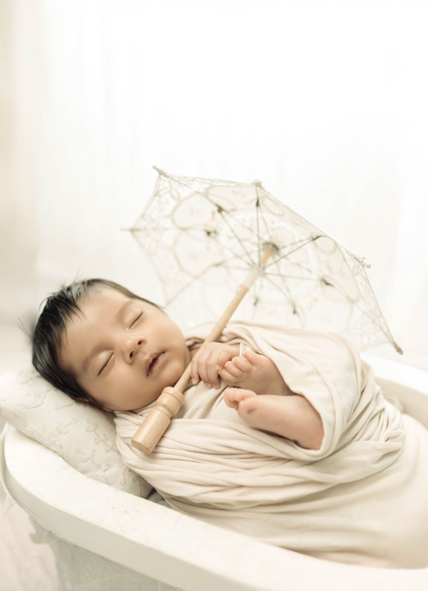 Peaceful newborn baby sleeping swaddled in cream wrap, holding a delicate lace parasol umbrella in a white bassinet.