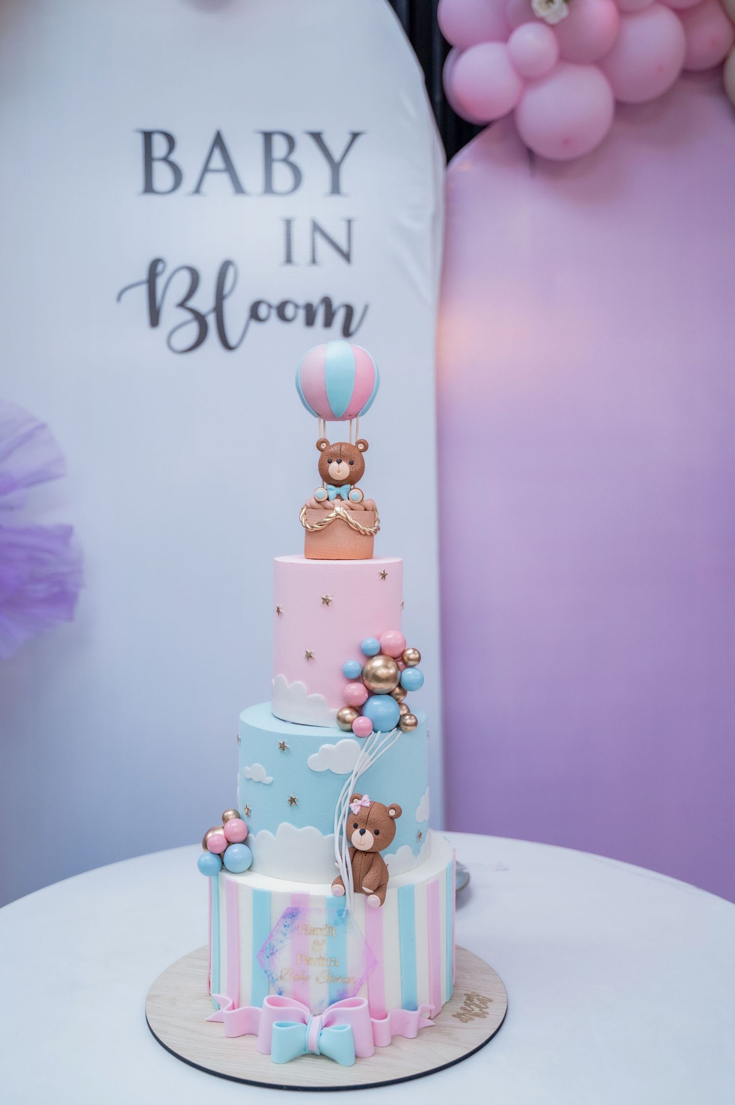 Three-tier baby shower cake with teddy bears and hot air balloon topper in pink and blue for a Baby in Bloom celebration.