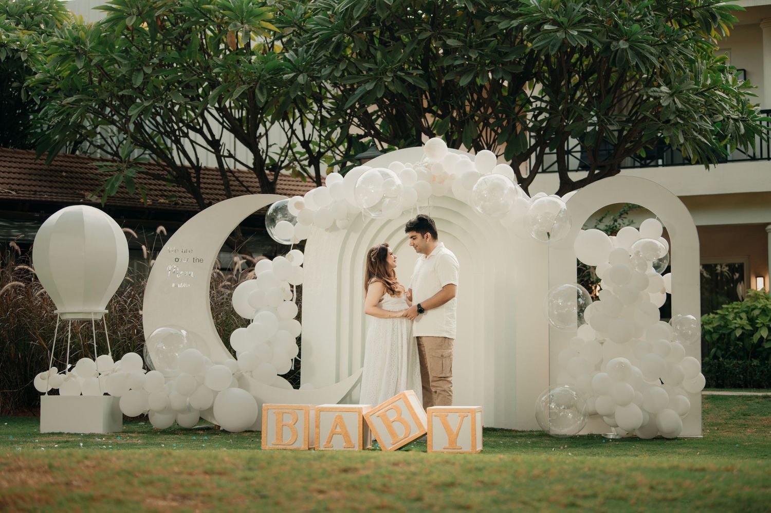Couple poses under white balloon arch with floral decor and BABY letter blocks at outdoor baby shower celebration.