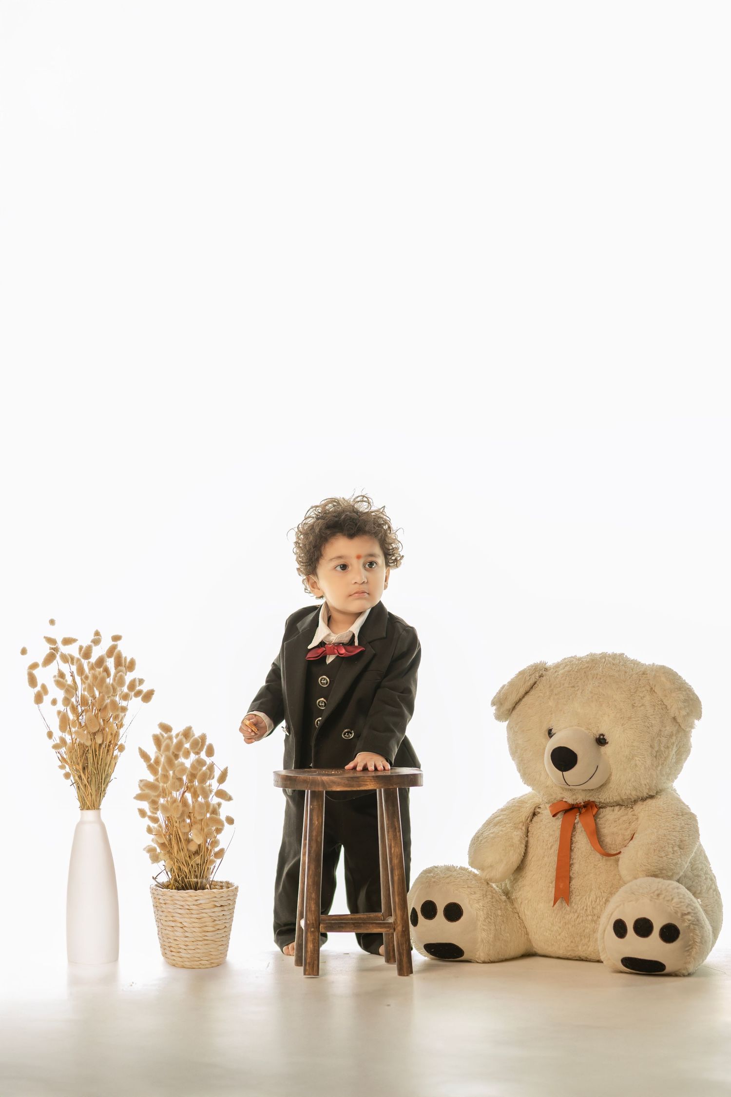 Toddler boy in black suit stands by wooden stool next to large teddy bear and dried flowers on white background.