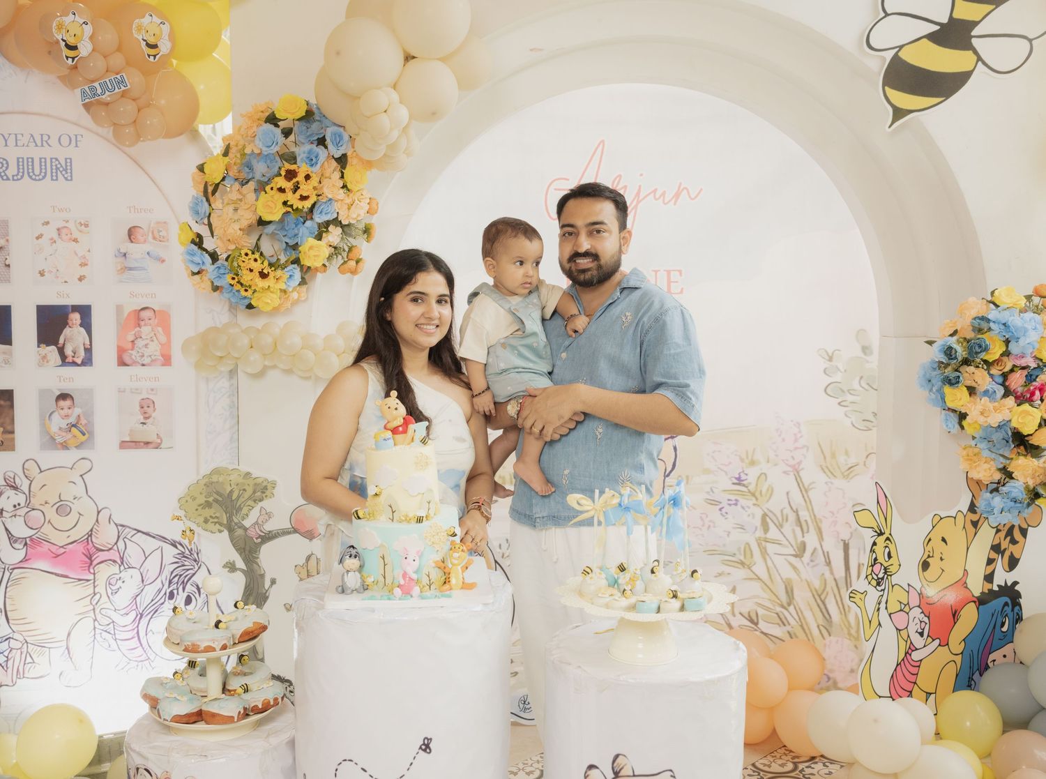 Family celebrating baby's first birthday with Winnie the Pooh themed cakes and decorations.