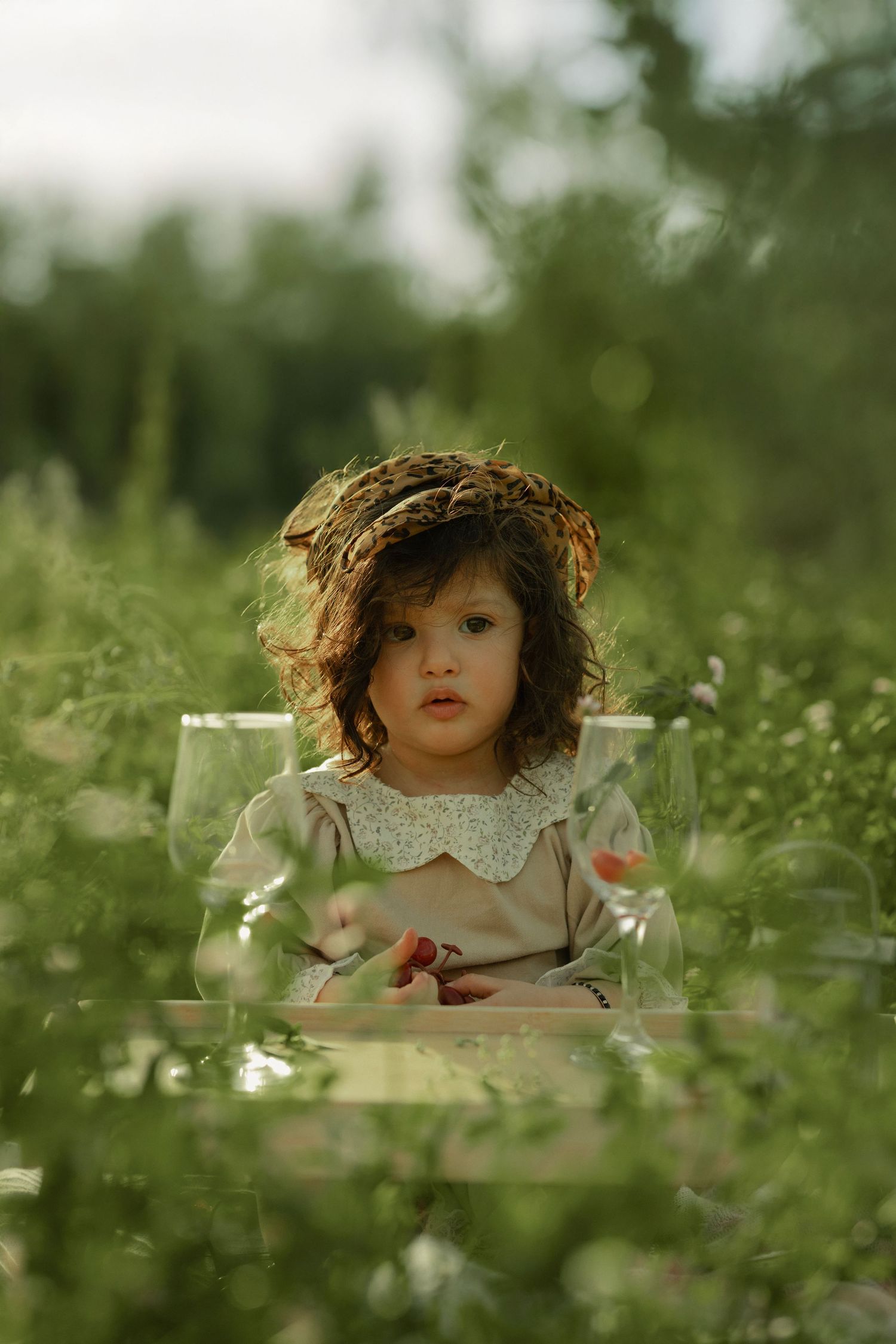 A young girl wearing a floral wreath crown sits in a chair surrounded by lush green meadow grass in soft natural light.