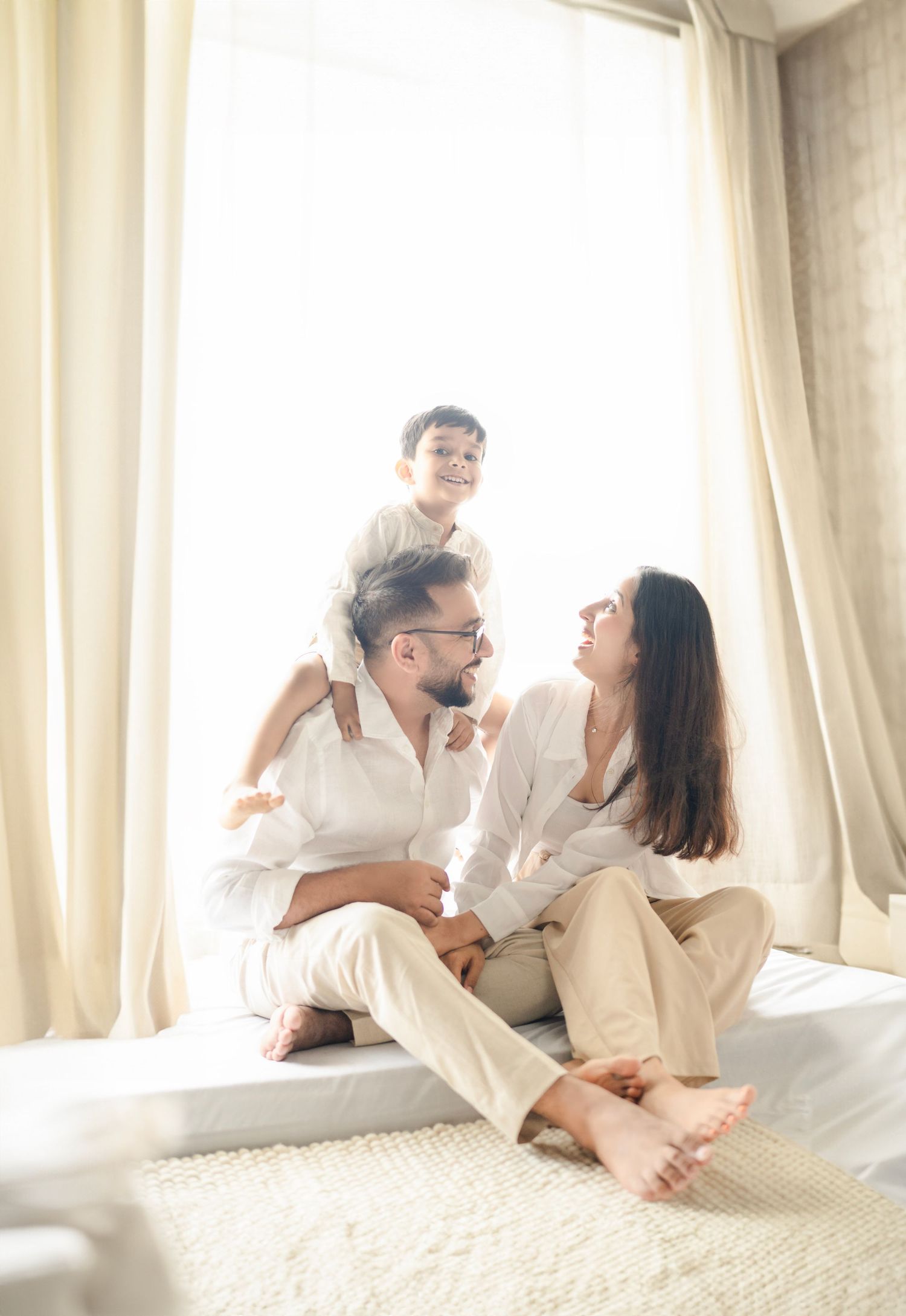 Happy family of three in white outfits sitting on a bed by a bright window, parents smiling at each other with child on shoulders.