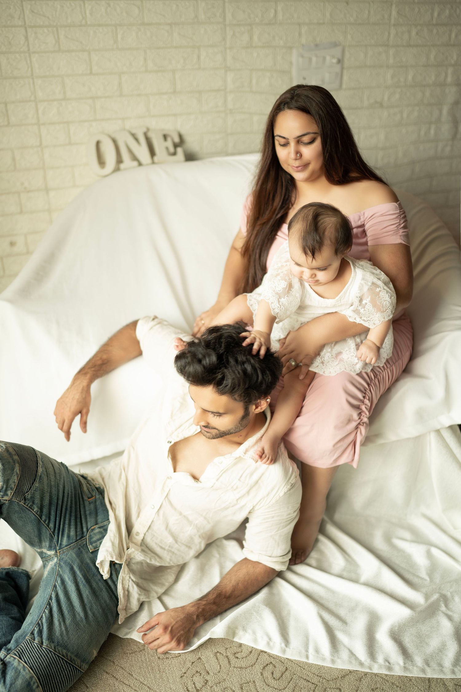 A young family with a baby girl relaxes on a white bed, with ONE letters on the wall, celebrating her first birthday.