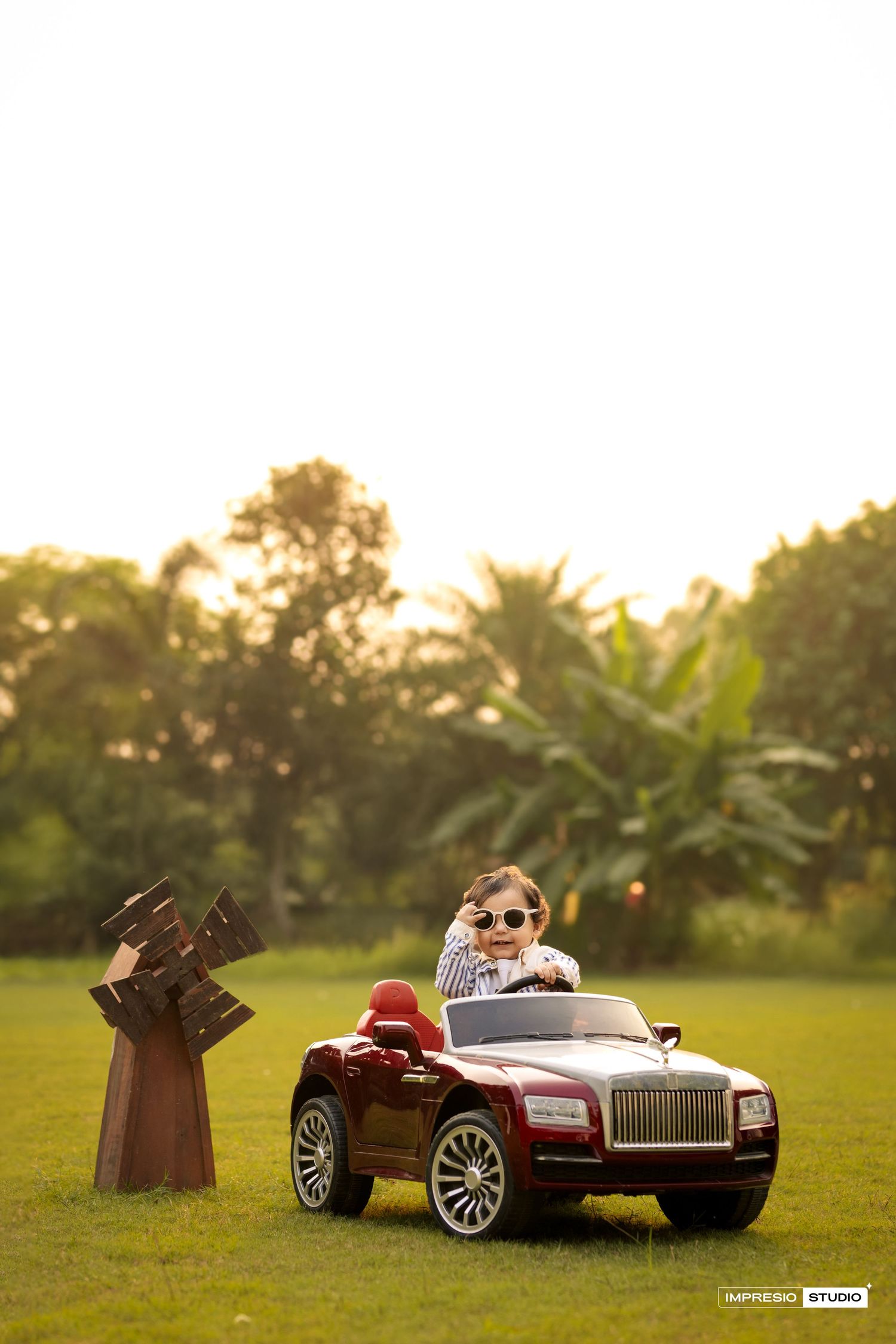 Child drives a red miniature luxury toy car on grass lawn at sunset with palm trees in background.