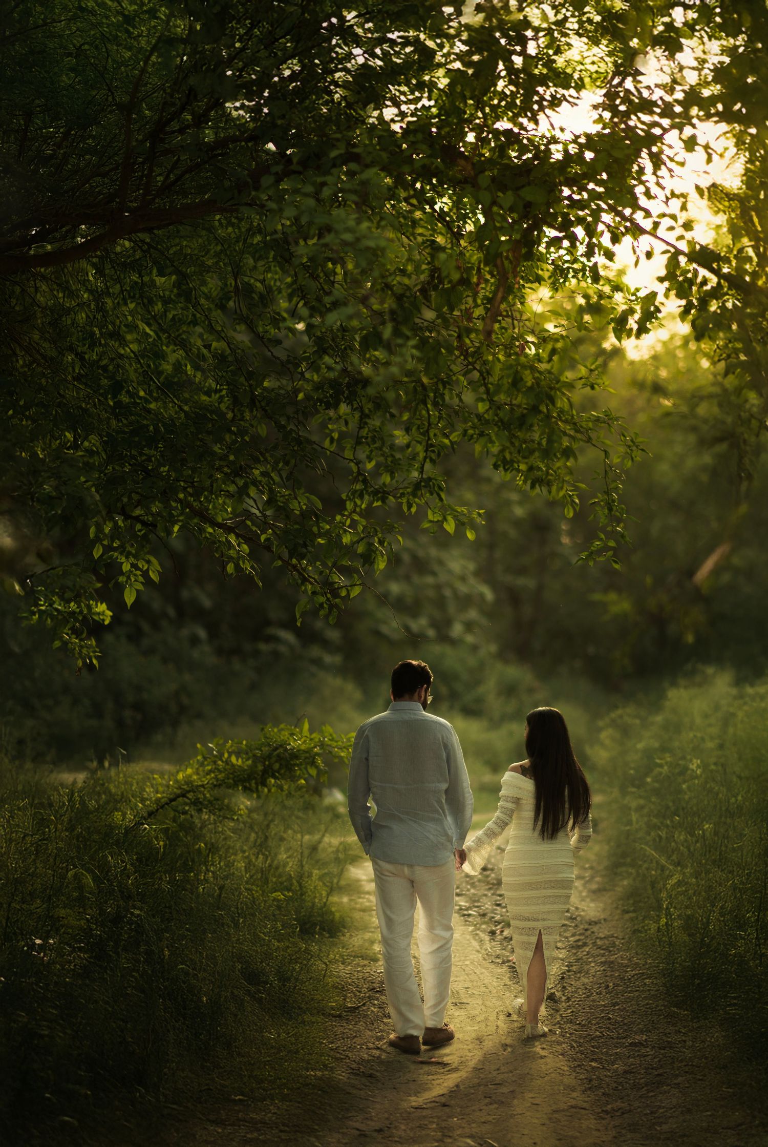 A couple walks hand in hand down a sunlit forest path during golden hour as sunbeams filter through green leaves.