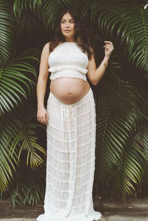 An expectant person poses in a white two-piece lace maternity dress against a backdrop of tropical palm fronds.