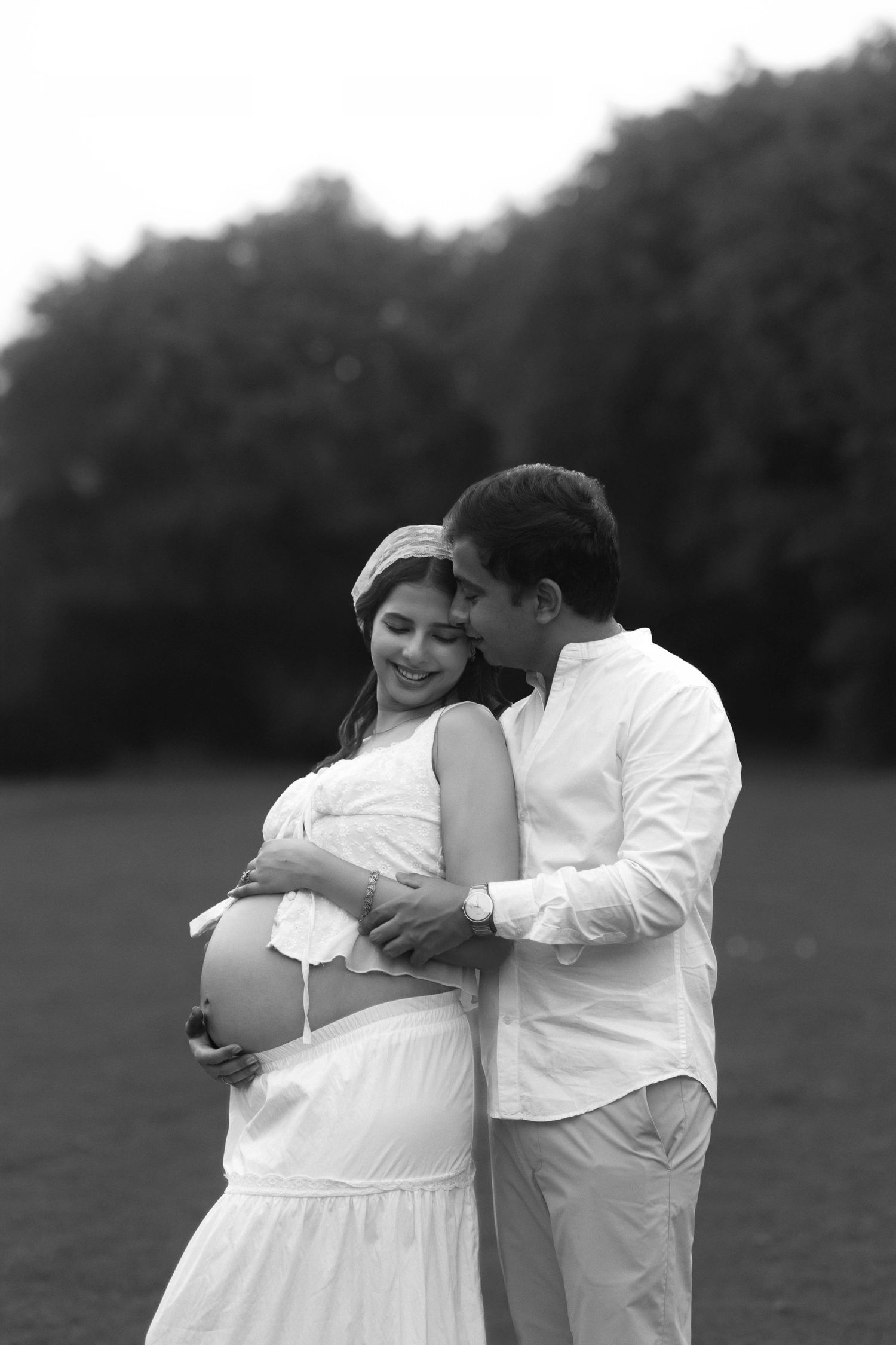 A romantic black and white maternity photo shows a couple embracing outdoors in white clothing.