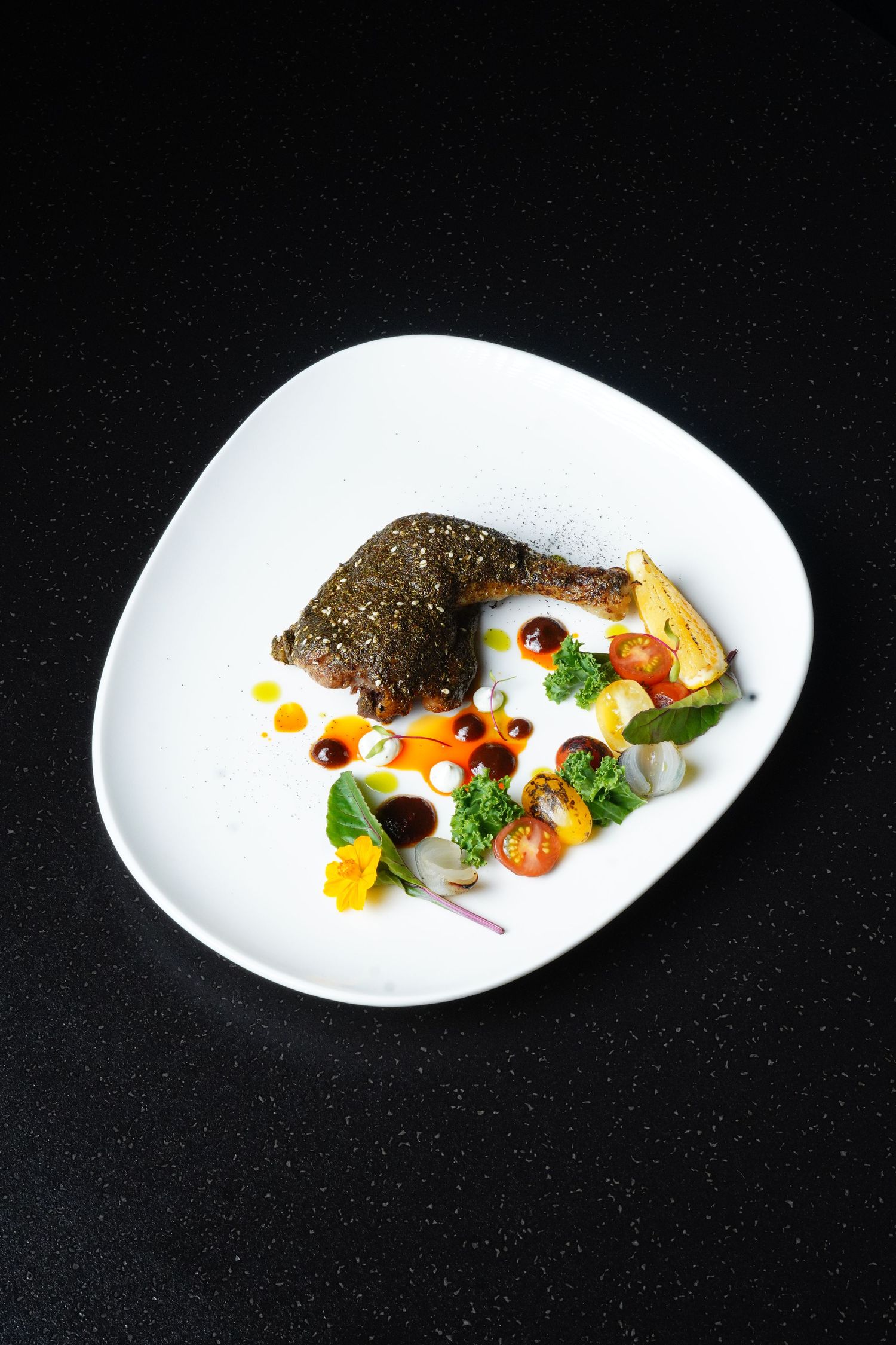 Elegantly plated fine dining dish featuring fish with colorful garnishes and sauces on a curved white plate against black.