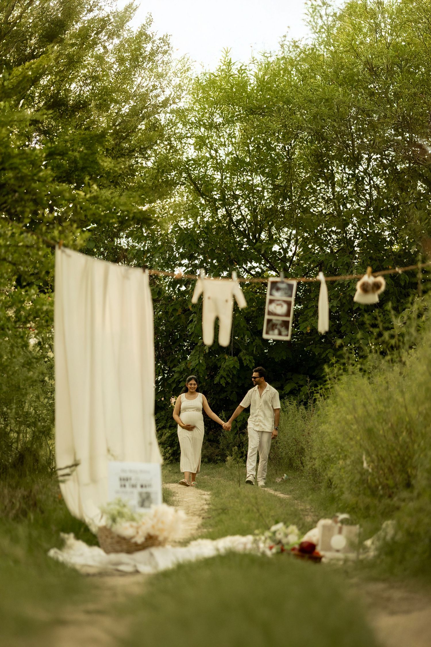 Pregnant couple holds hands outdoors near clothesline with baby onesie and ultrasound photo, surrounded by lush greenery.