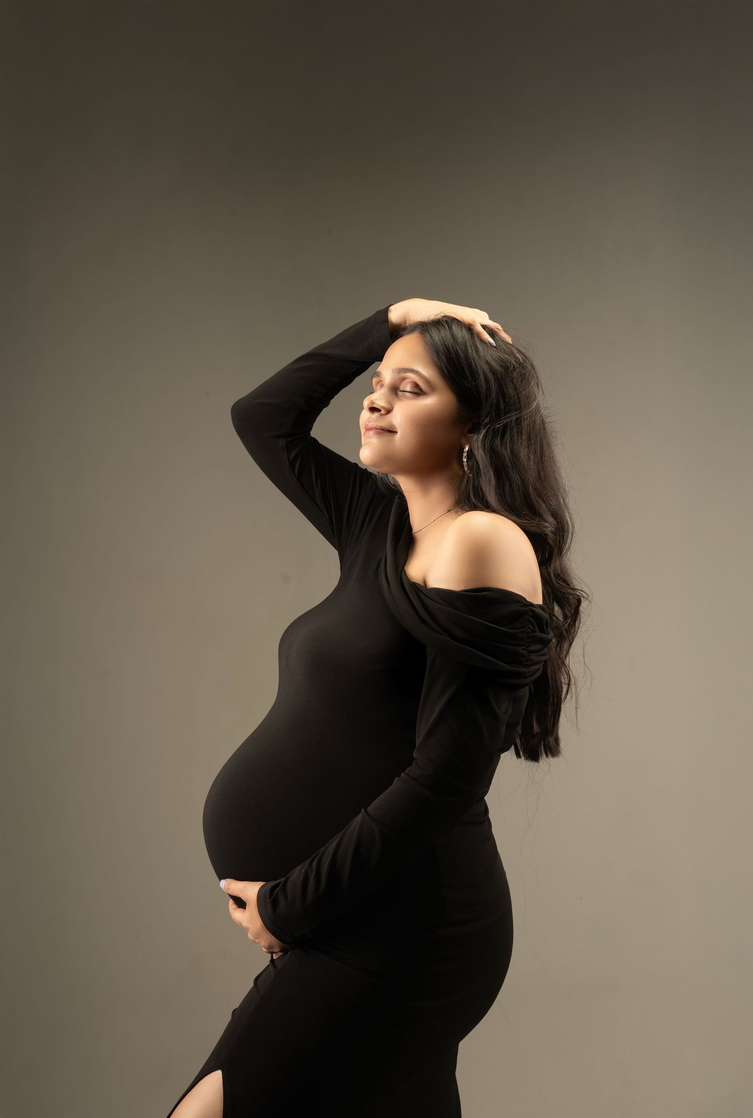 Pregnant woman in black off-shoulder dress posing gracefully, cradling baby bump in professional maternity photo shoot.