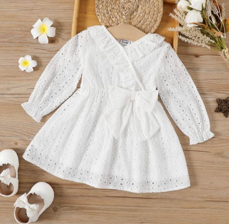 White eyelet lace baby dress with long sleeves displayed with floral accents and baby shoes on wooden surface.