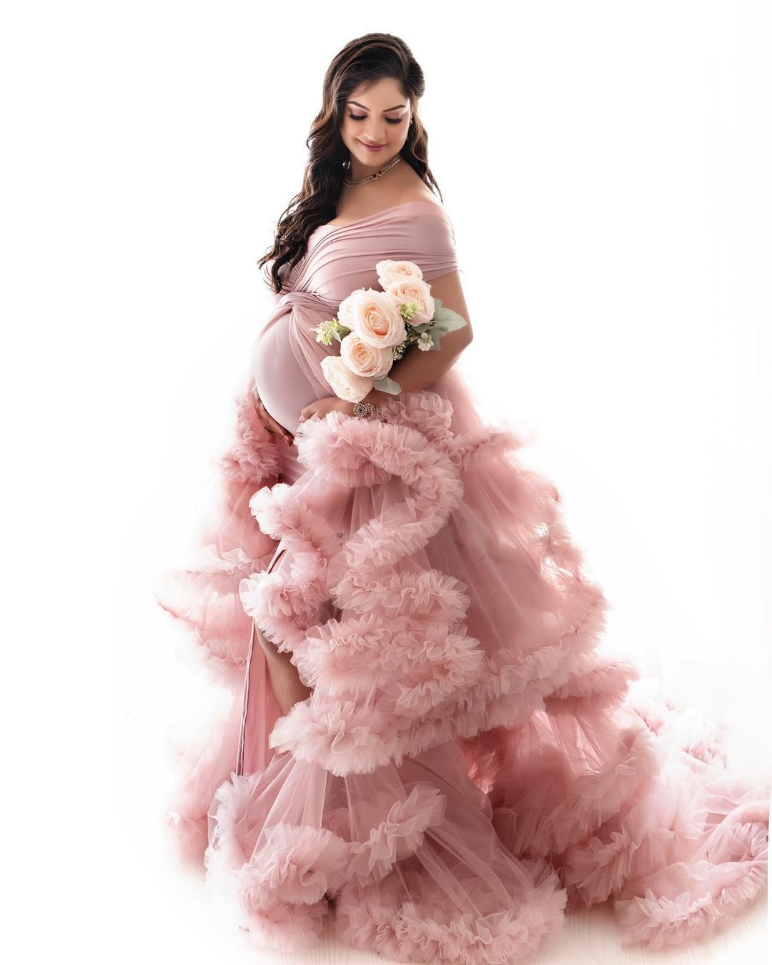 A feminine maternity portrait in a pink ruffled gown holding peach roses.