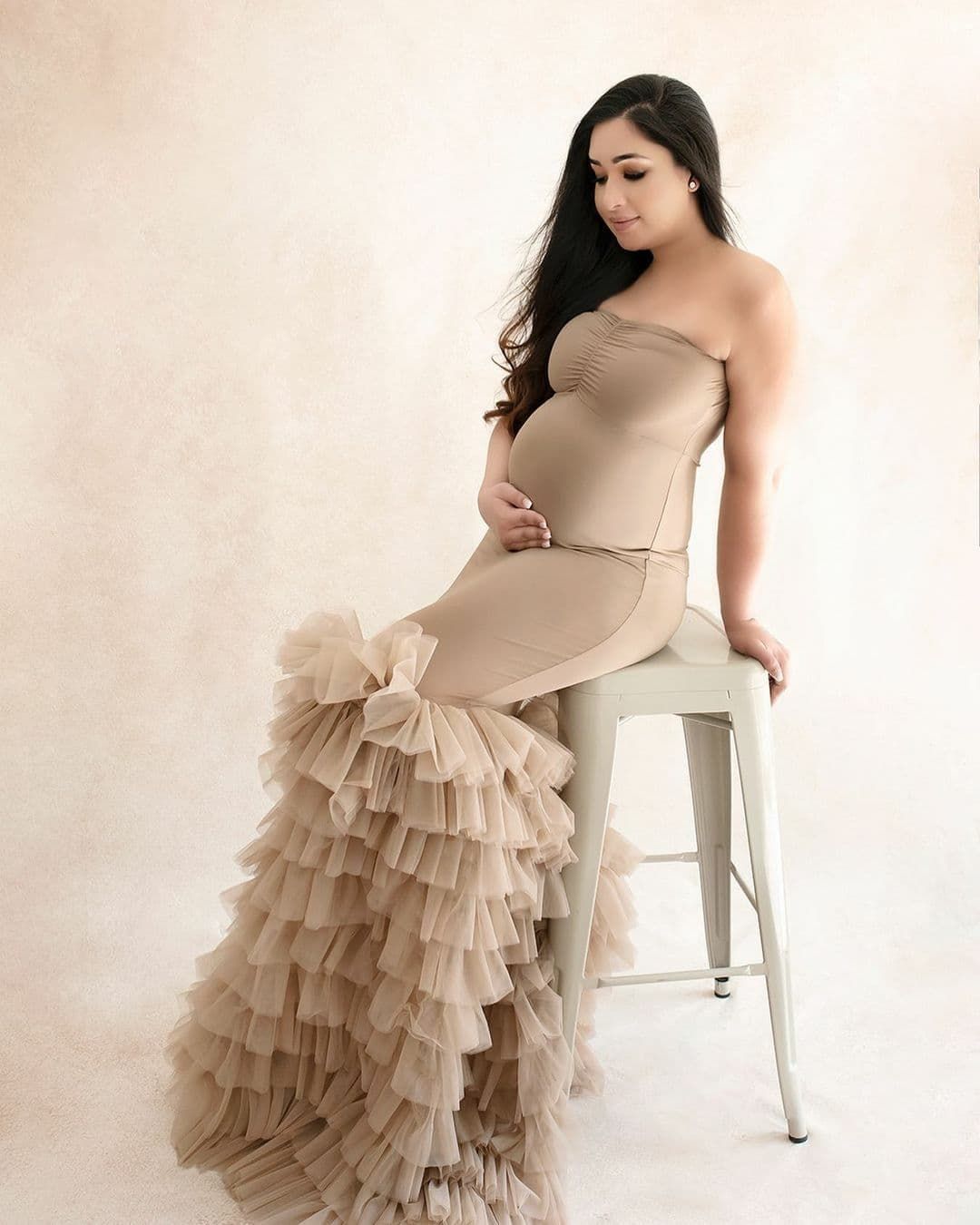 Elegant maternity photo featuring a beige strapless gown with dramatic ruffled tulle layers sitting on a metal stool.