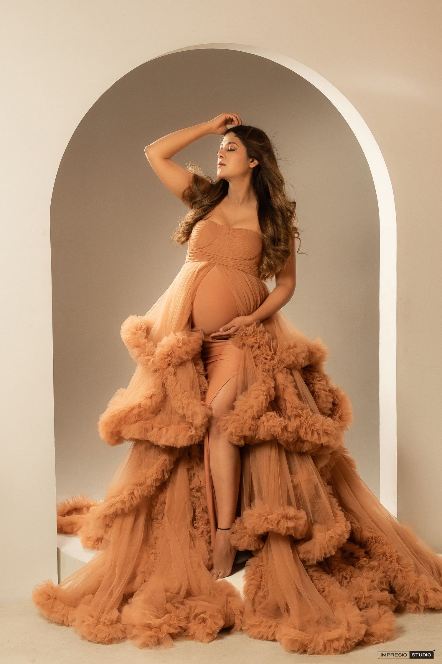 Maternity photoshoot featuring a dramatic peach-colored tulle gown with ruffled layers in an arched setting.
