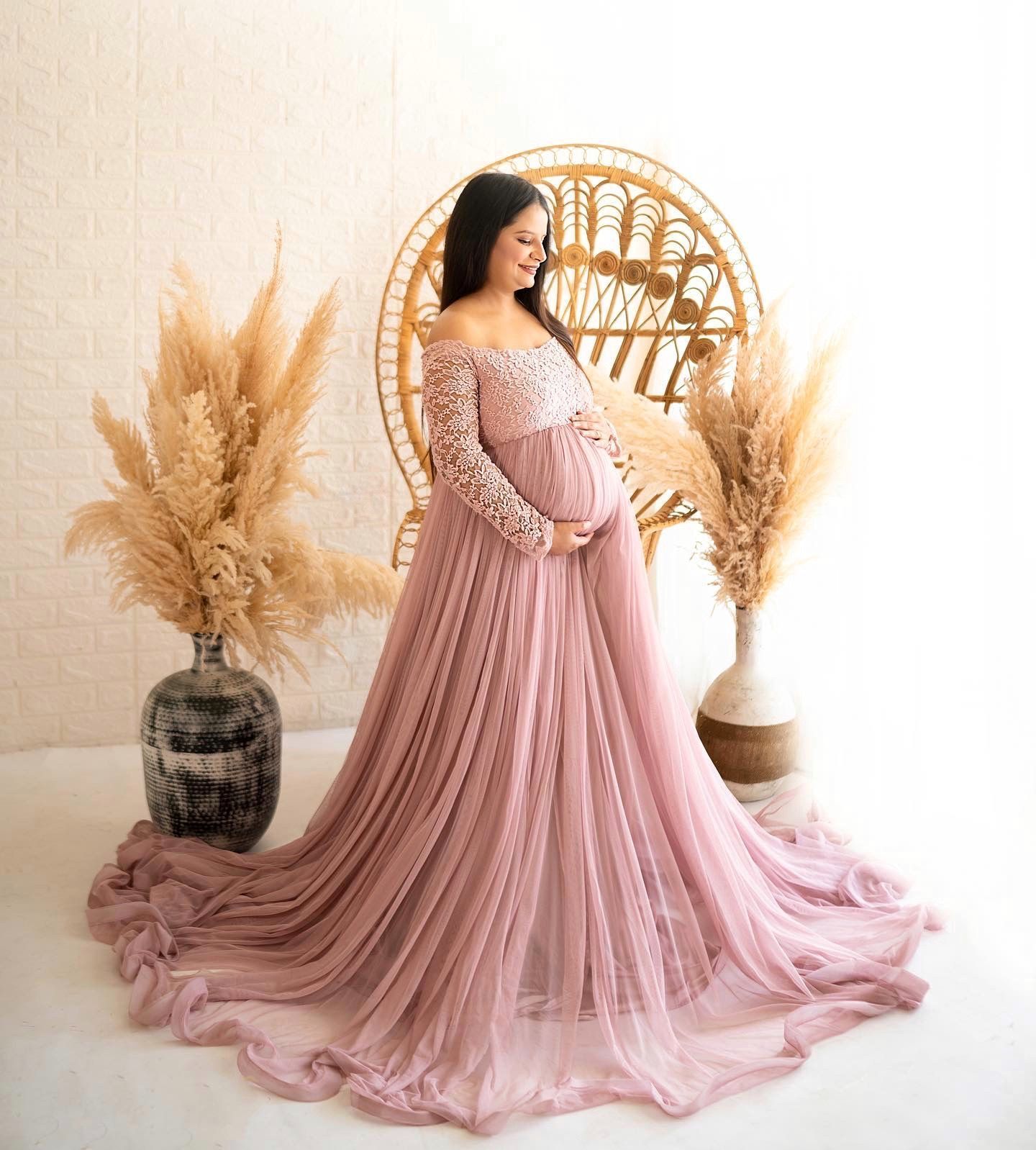 A flowing pink maternity dress is photographed with decorative pampas grass on a light beige background.