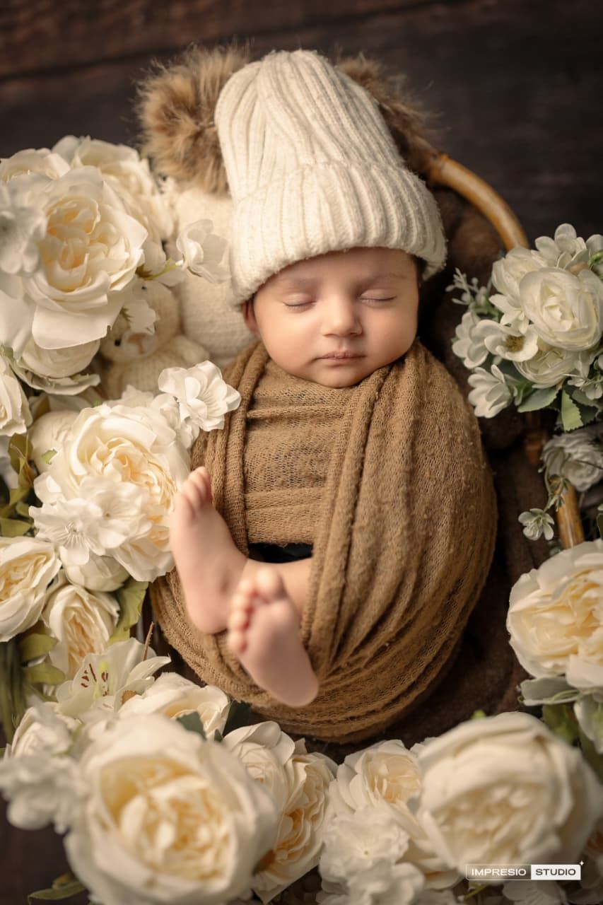 Newborn baby swaddled in brown wrap and white knit hat, sleeping peacefully in a basket surrounded by cream roses.