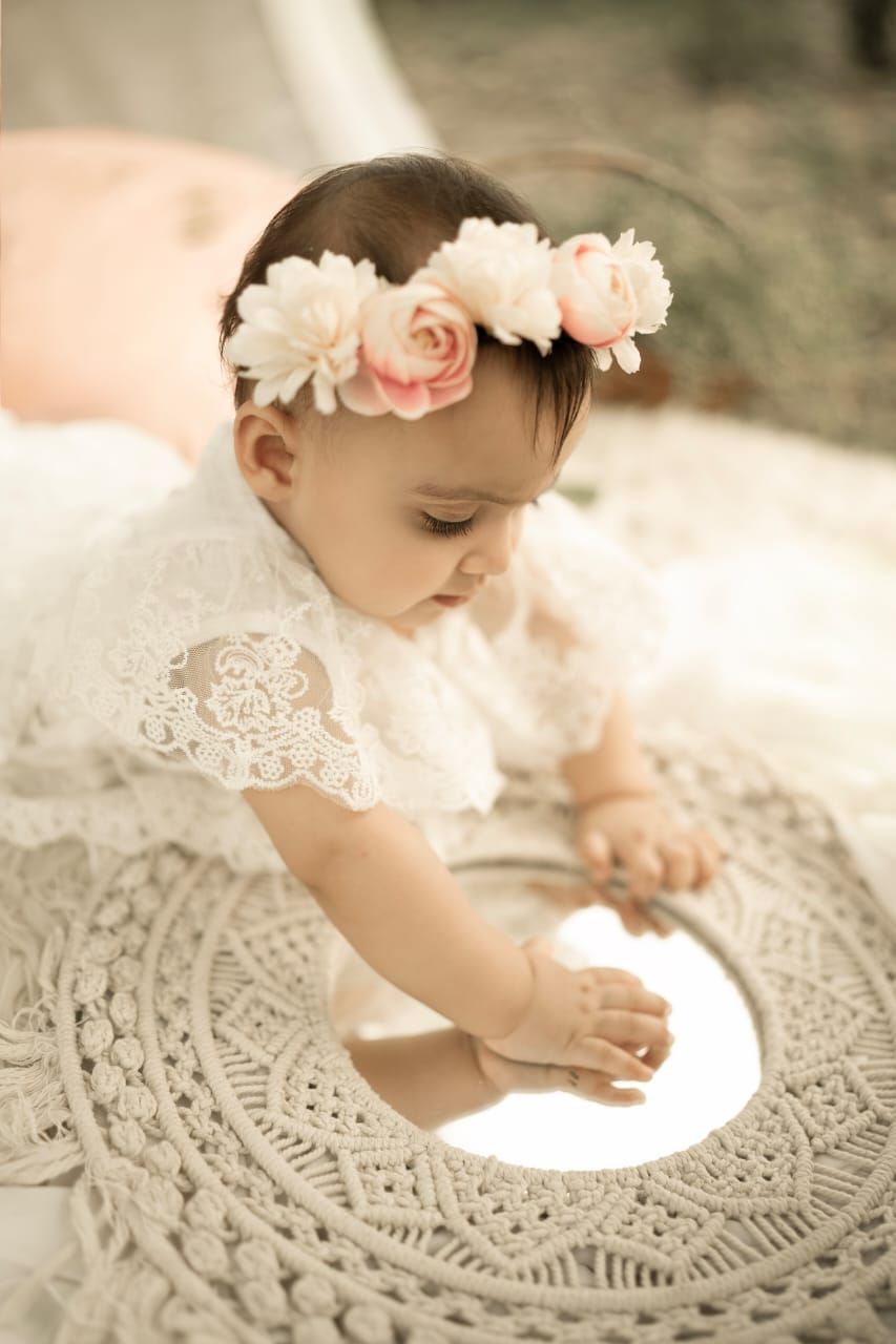 Baby girl in white lace dress and floral crown gazes at mirror on crochet mat in soft outdoor setting.