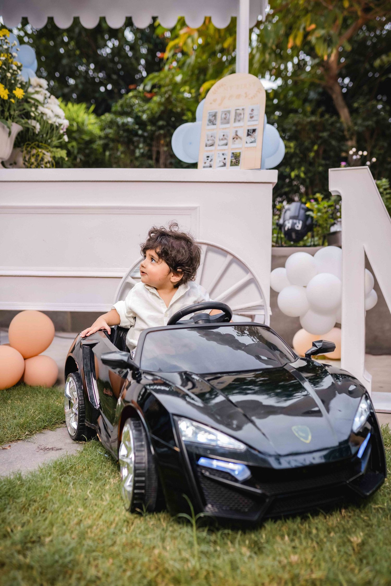 A toddler sits in a black ride-on toy sports car outdoors at a birthday party with balloons and decorations.