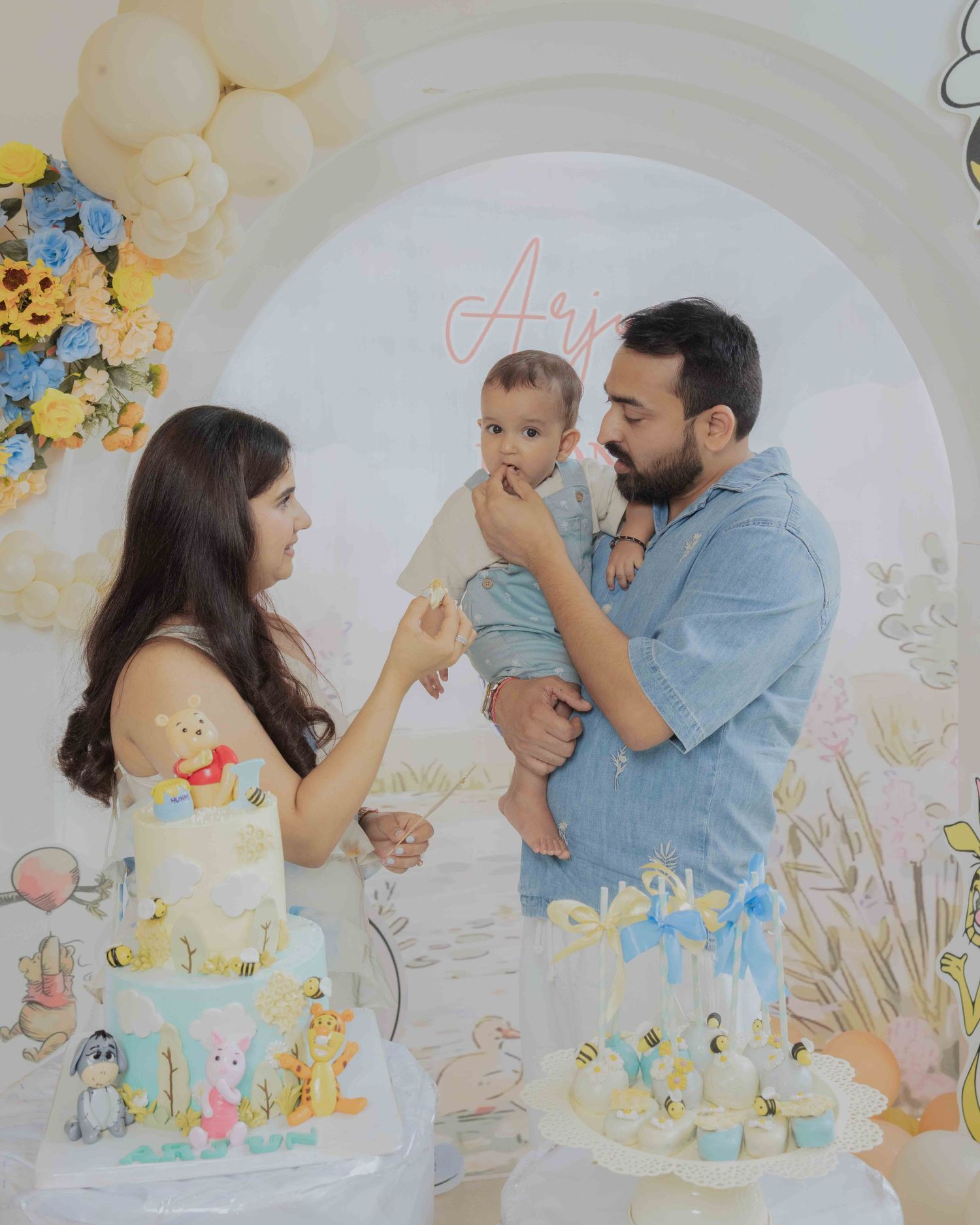 Family celebrating baby's first birthday with a decorated cake and balloon arch backdrop.