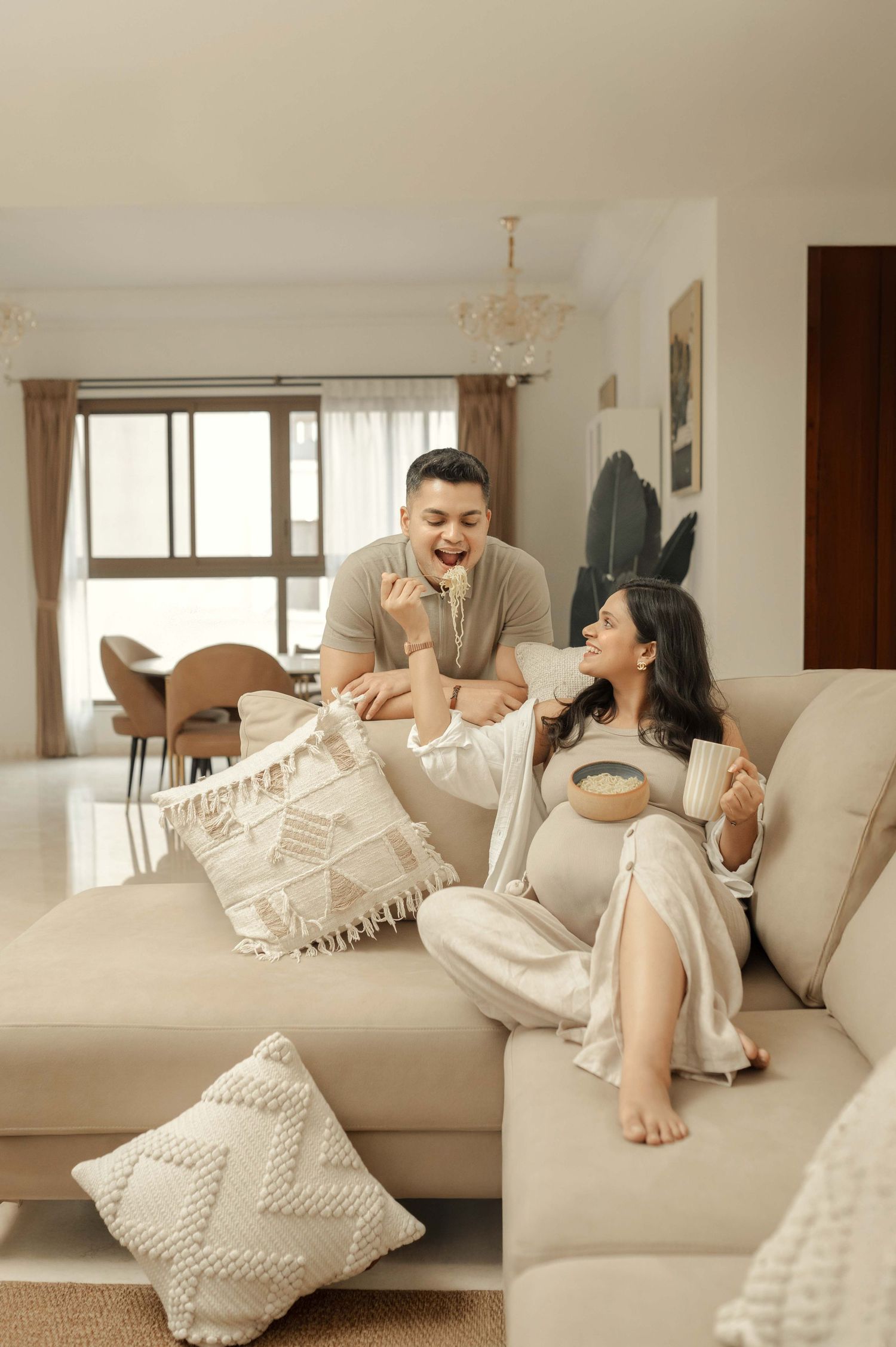 Happy couple relaxing on a beige sofa, sharing snacks and laughing together in a bright, modern living room.