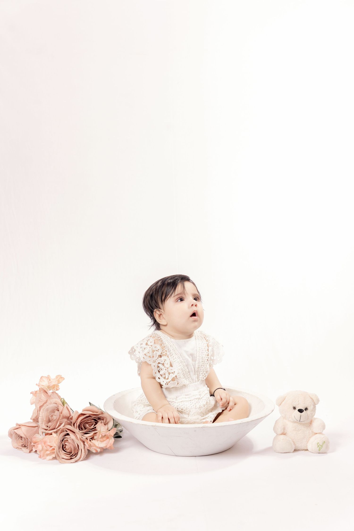 Baby girl in white lace dress sitting in a bowl with roses and teddy bear on white background.