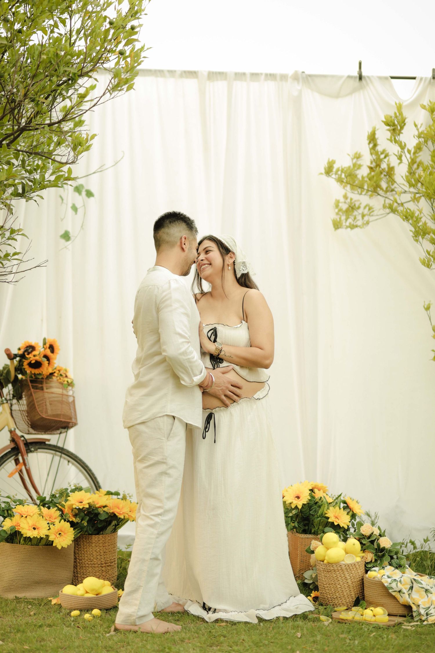 A couple in wedding attire embraces outdoors, surrounded by sunflowers and yellow flowers against a white curtain backdrop.