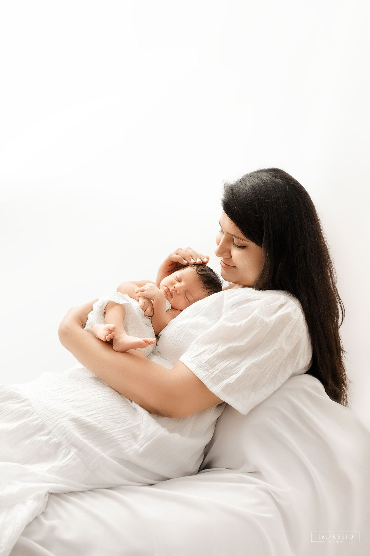 A mother with dark hair tenderly holds her sleeping newborn baby while resting in white bedding.