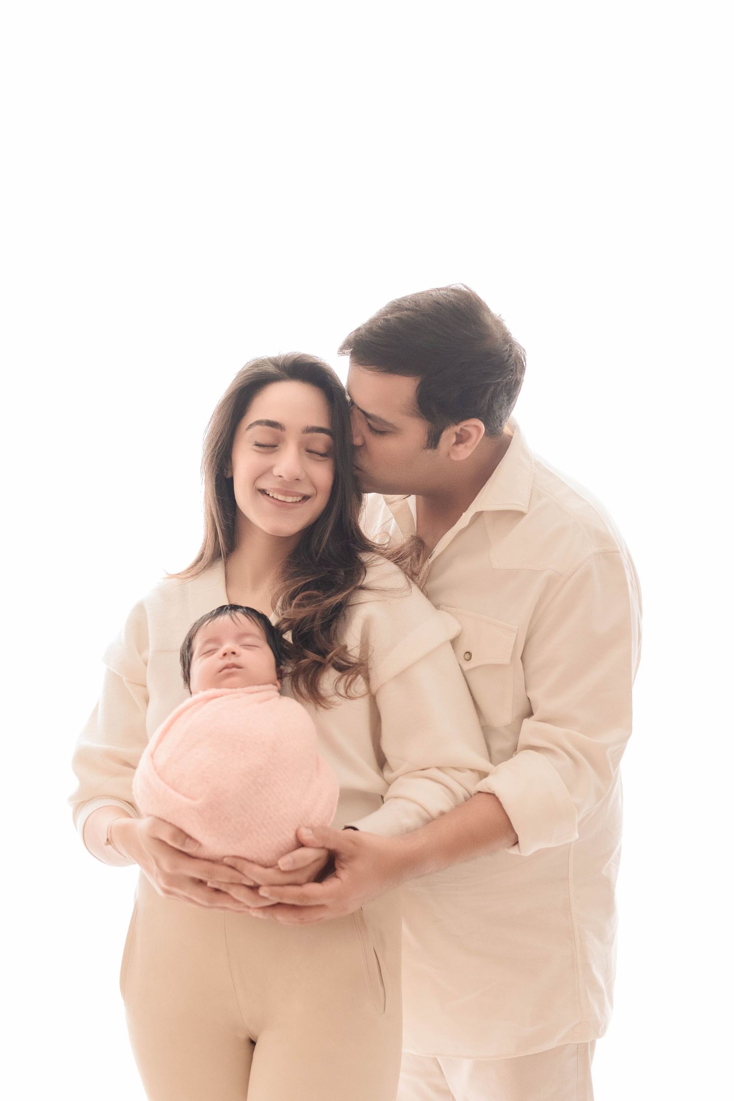 A happy couple in beige outfits holds their sleeping newborn wrapped in pink against a white background.