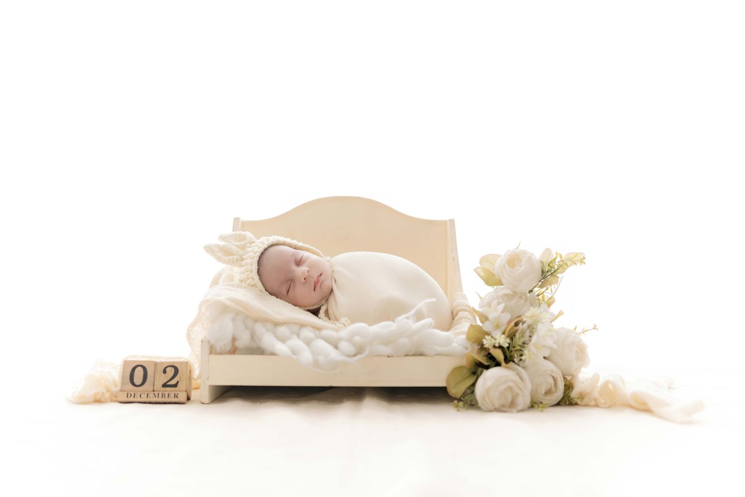 Newborn baby sleeping on miniature cream bed with white flowers and date blocks showing 02 in studio setting.