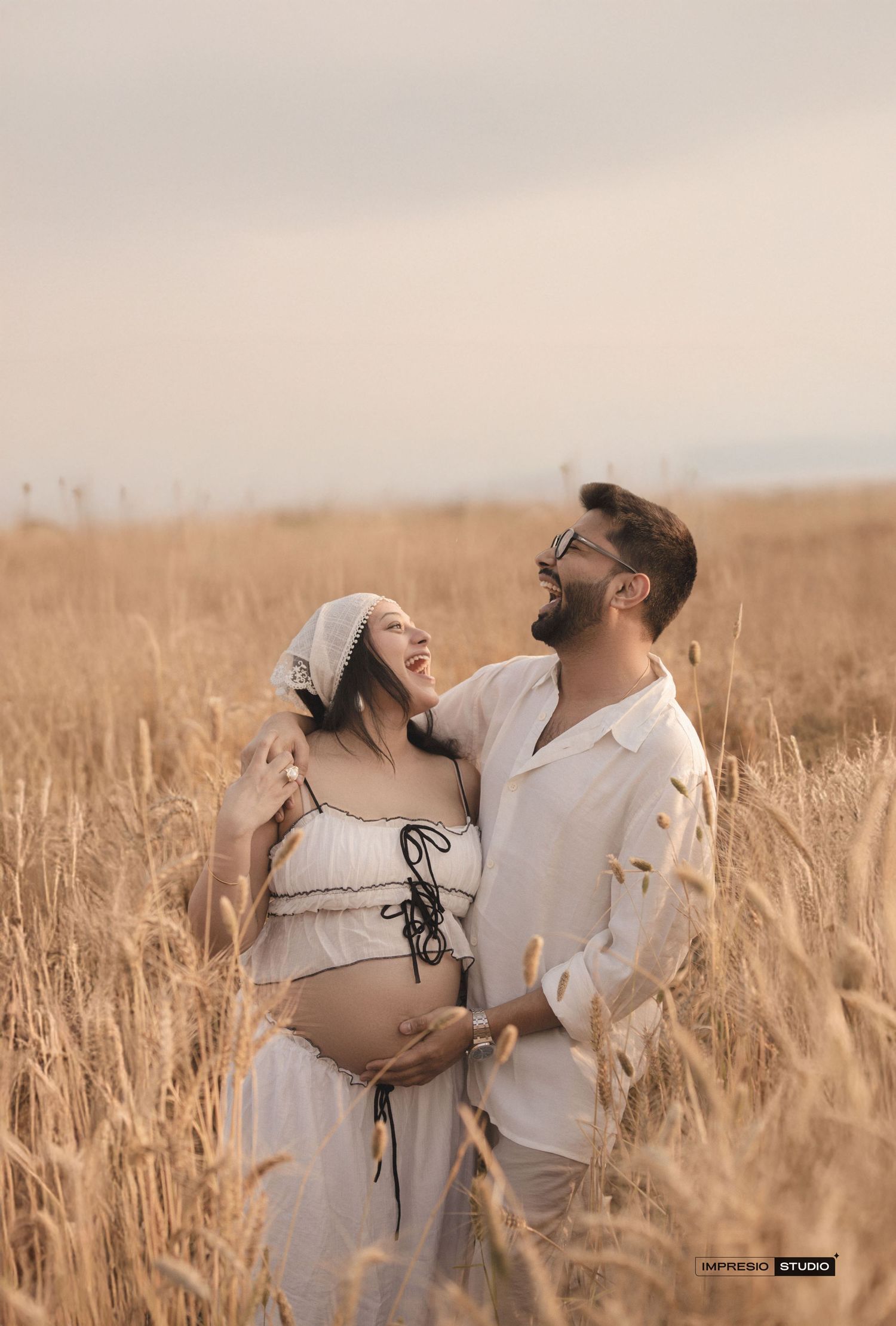 Couple in white outfits sharing a tender moment in a golden wheat field during a maternity photoshoot.