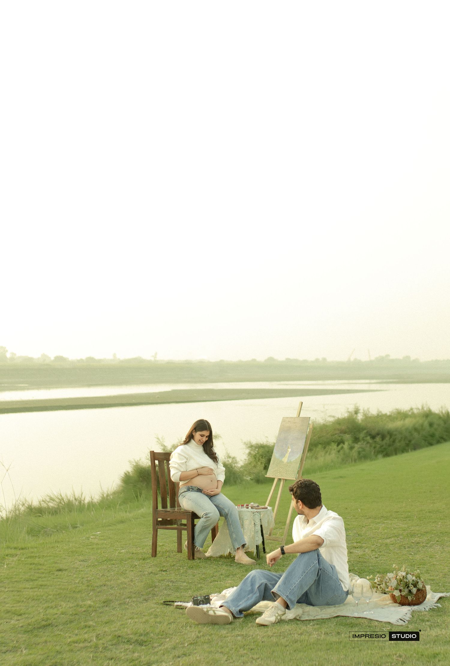Pregnant woman sitting on chair outdoors by lake while man paints her portrait on easel during maternity photoshoot.