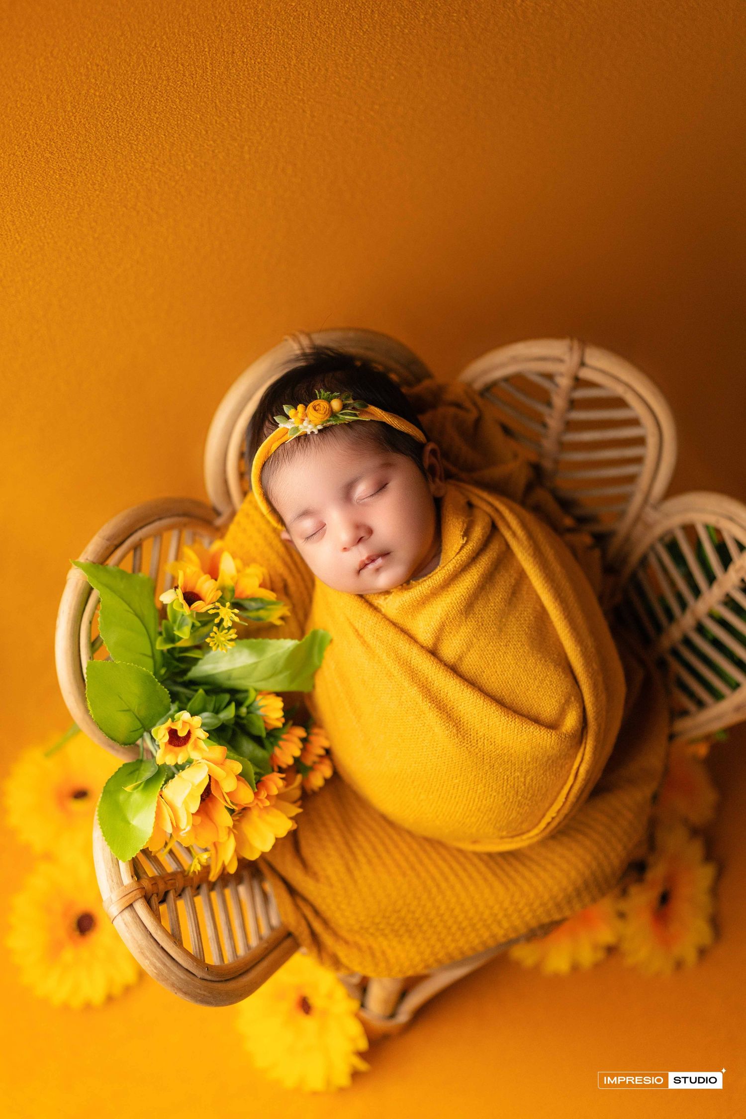 Newborn baby wrapped in yellow swaddle sleeping in rattan basket surrounded by sunflowers.