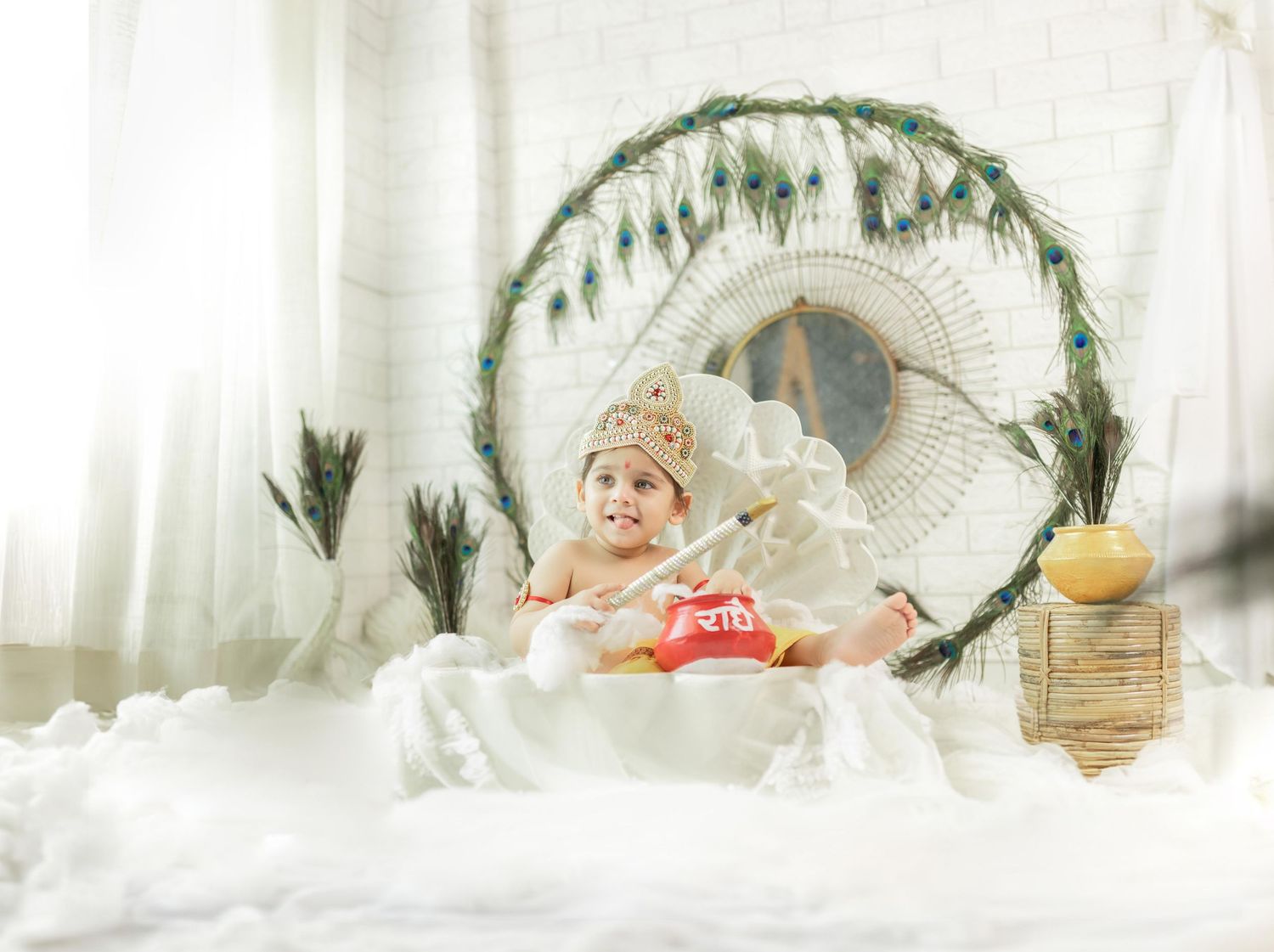 Baby in crown sits in boho peacock chair surrounded by white fluff and greenery.