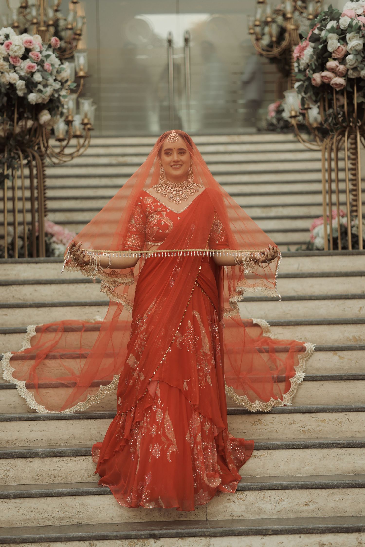 Bride in red lehenga with orange dupatta poses on marble stairs at Indian wedding ceremony.
