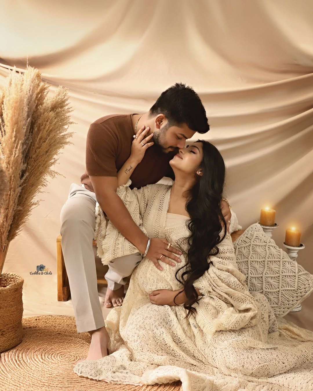 Romantic couple pose with candlelit boho maternity decor and pampas grass against draped fabric.