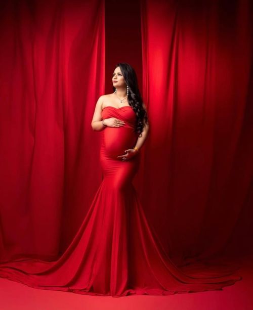 Stunning maternity photo featuring a flowing red gown against dramatic red curtains in elegant studio lighting.