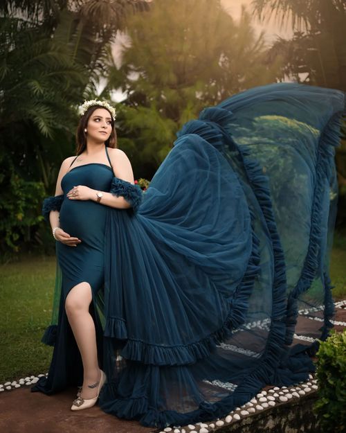 Flowing navy blue maternity gown billowing dramatically in outdoor garden setting with palm trees at sunset.