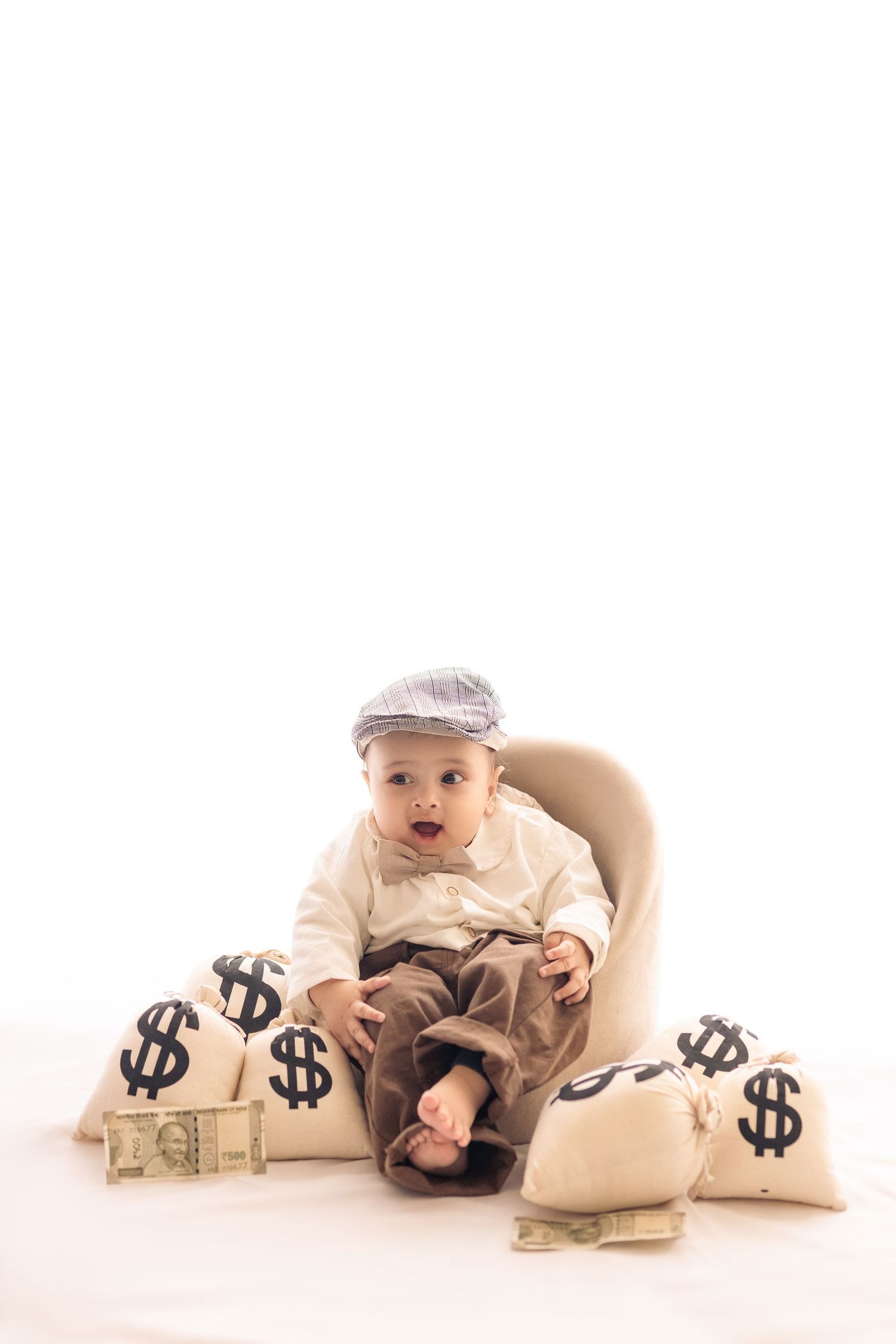 Small baby in beige outfit lying on floor with decorative money bags featuring dollar signs.