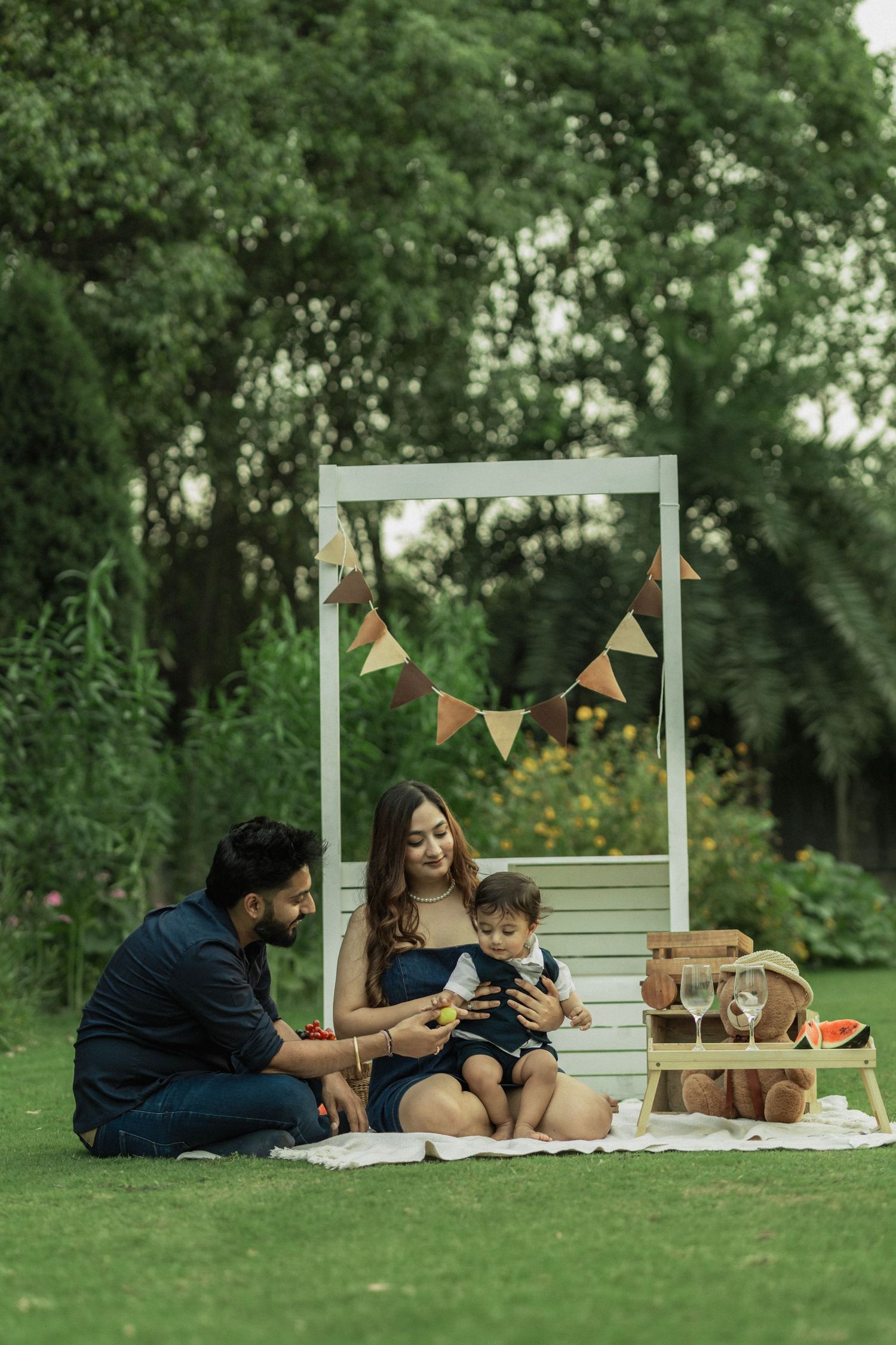 Young family with baby enjoying a picnic on green grass in a garden with bunting decorations and wooden backdrop.