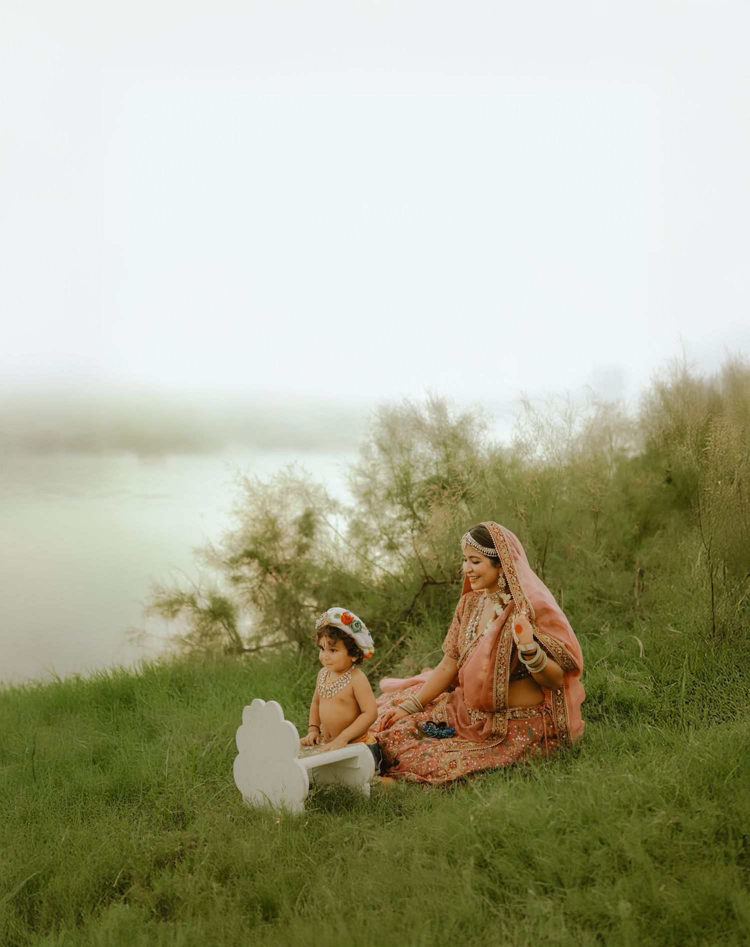 A bride in traditional Indian attire sits on grass with a child near a serene lakeside, surrounded by lush greenery.