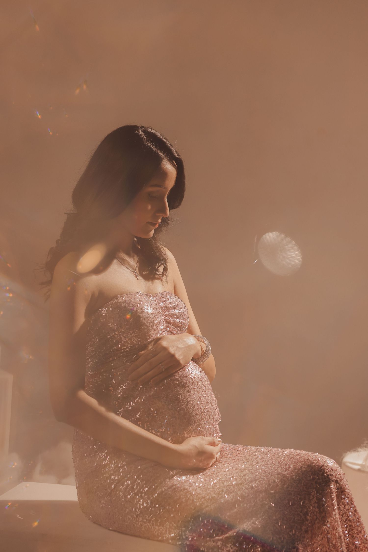 Pregnant woman in a shimmering pink sequin gown cradling her baby bump in a dreamy, glowing studio setting.