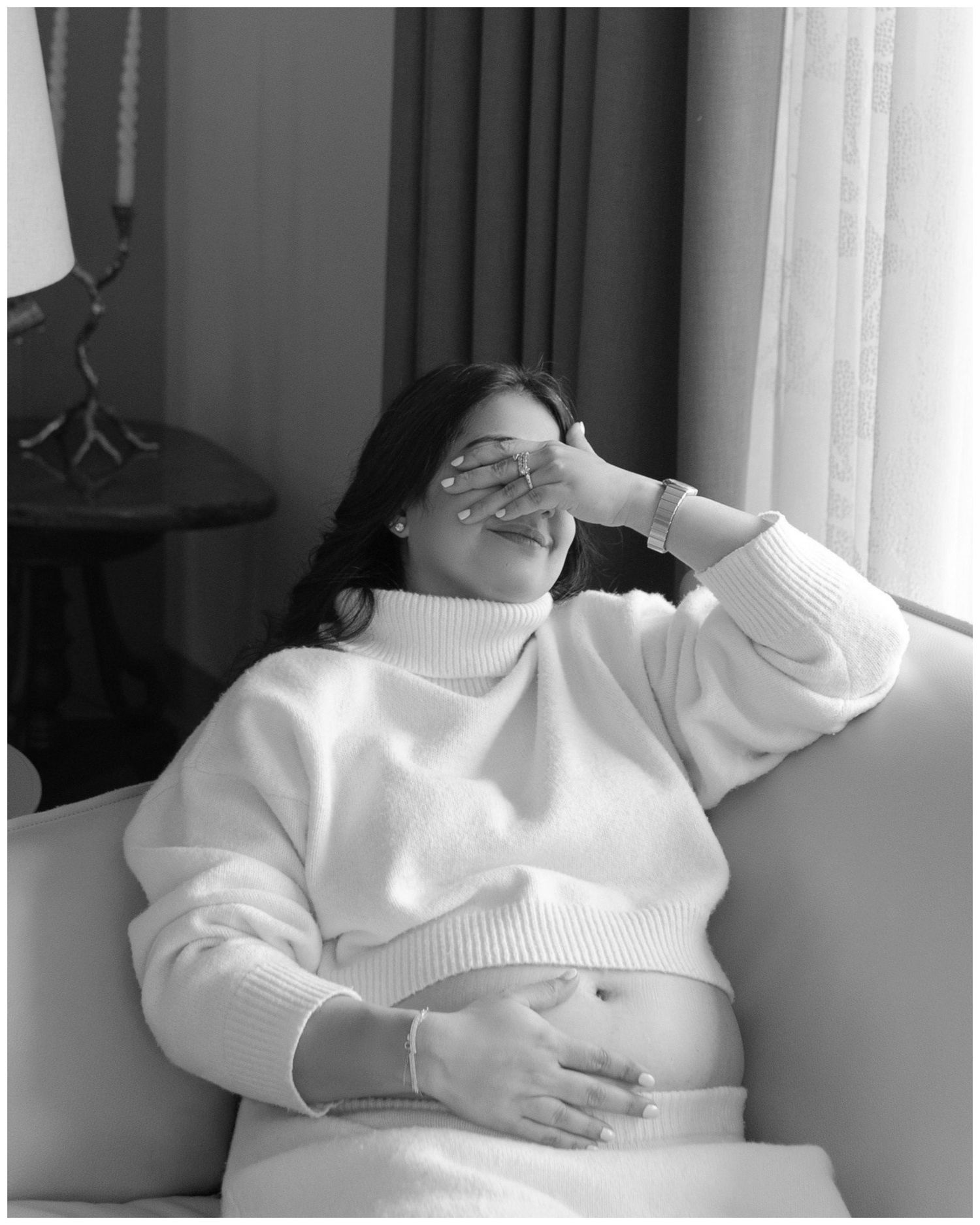 Black and white photo of a pregnant woman in a white turtleneck sweater sitting on a sofa, laughing.