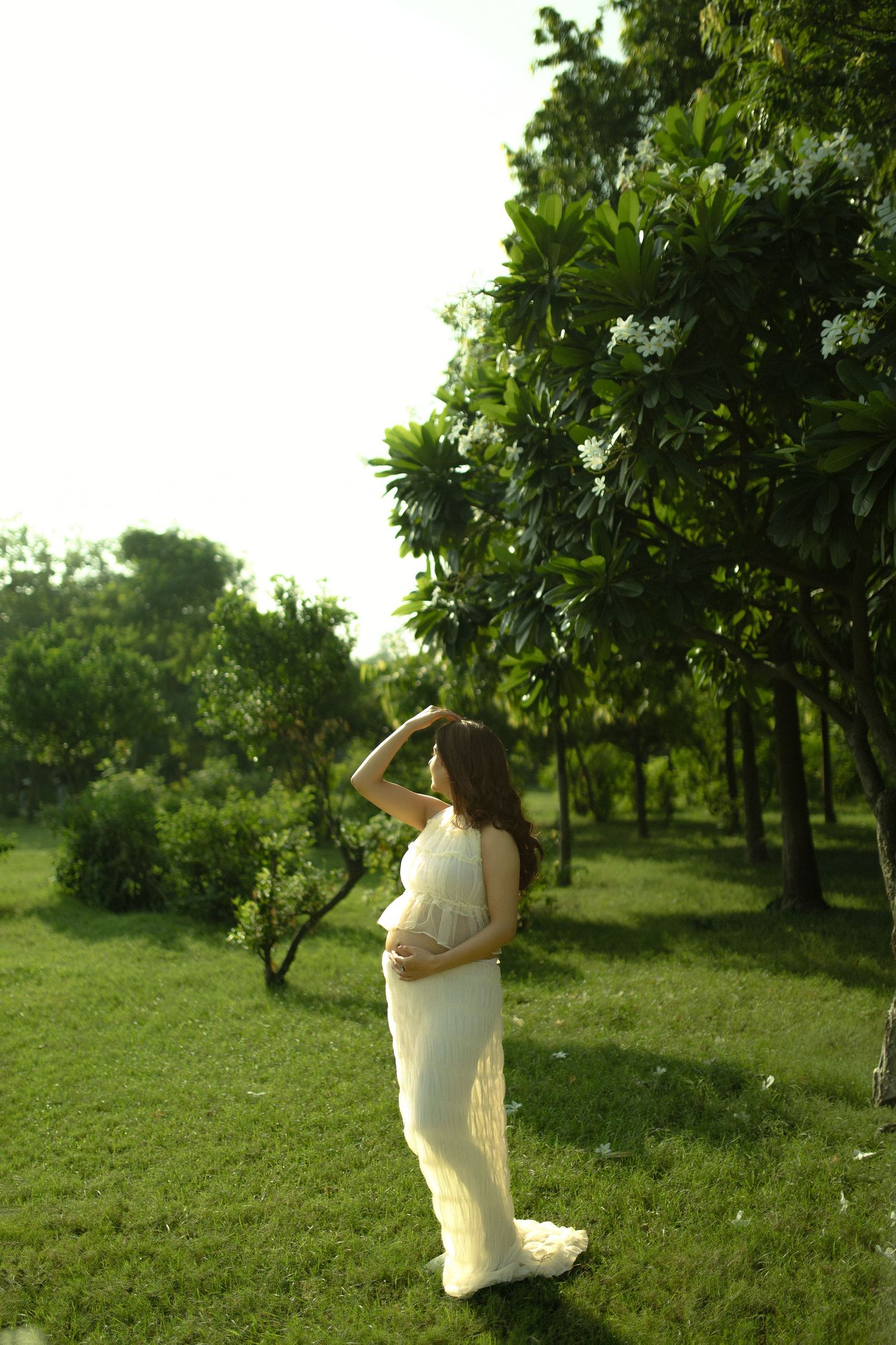 A person in a flowing white dress poses gracefully in a lush green garden setting with trees in the background.